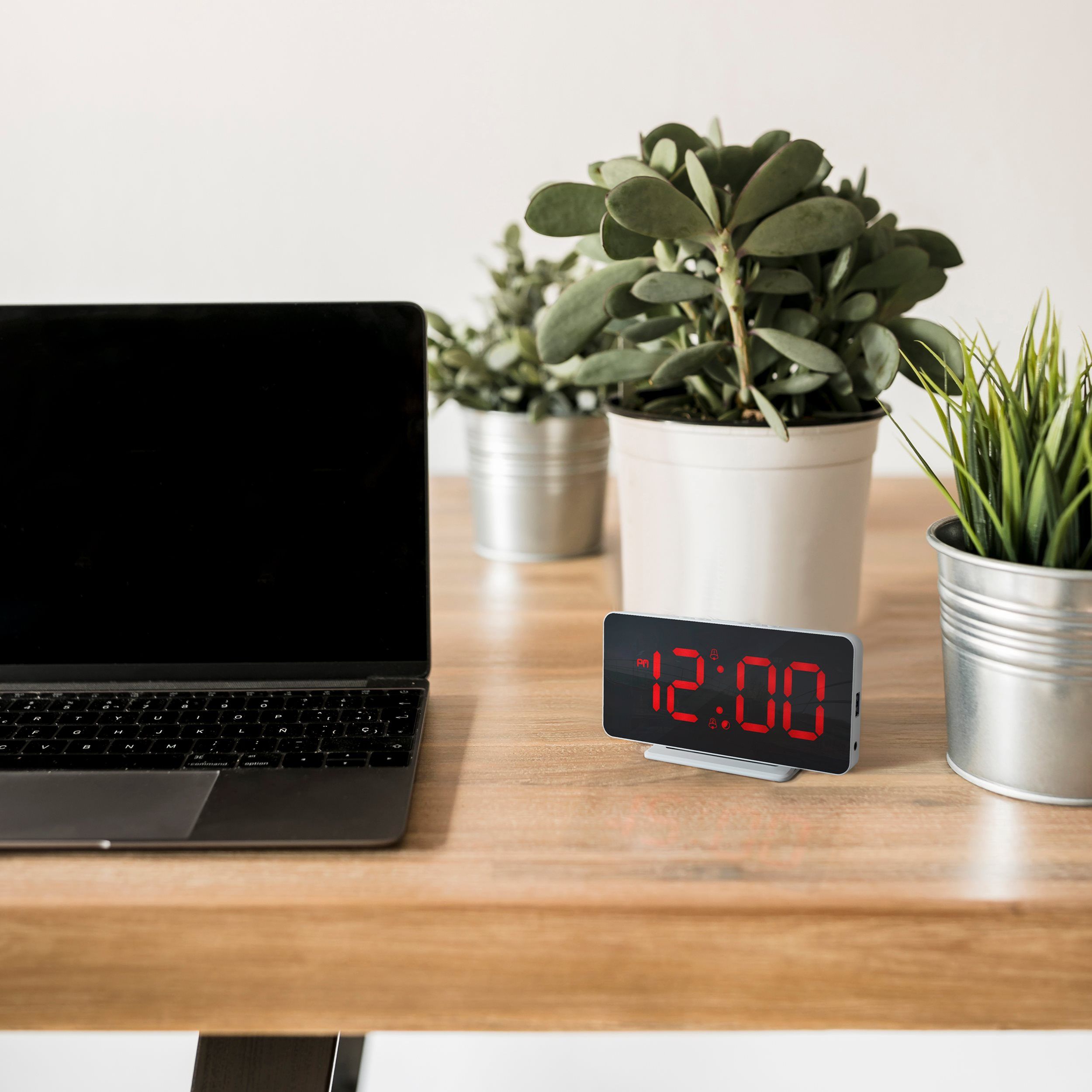 Laptop, clock, and plants on a wooden desk against a white wall.