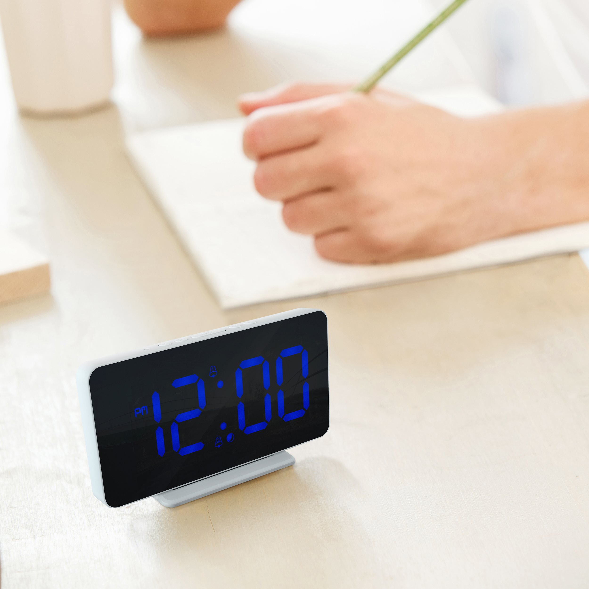 Digital clock showing 12:00 am on a table, person writing in background.
