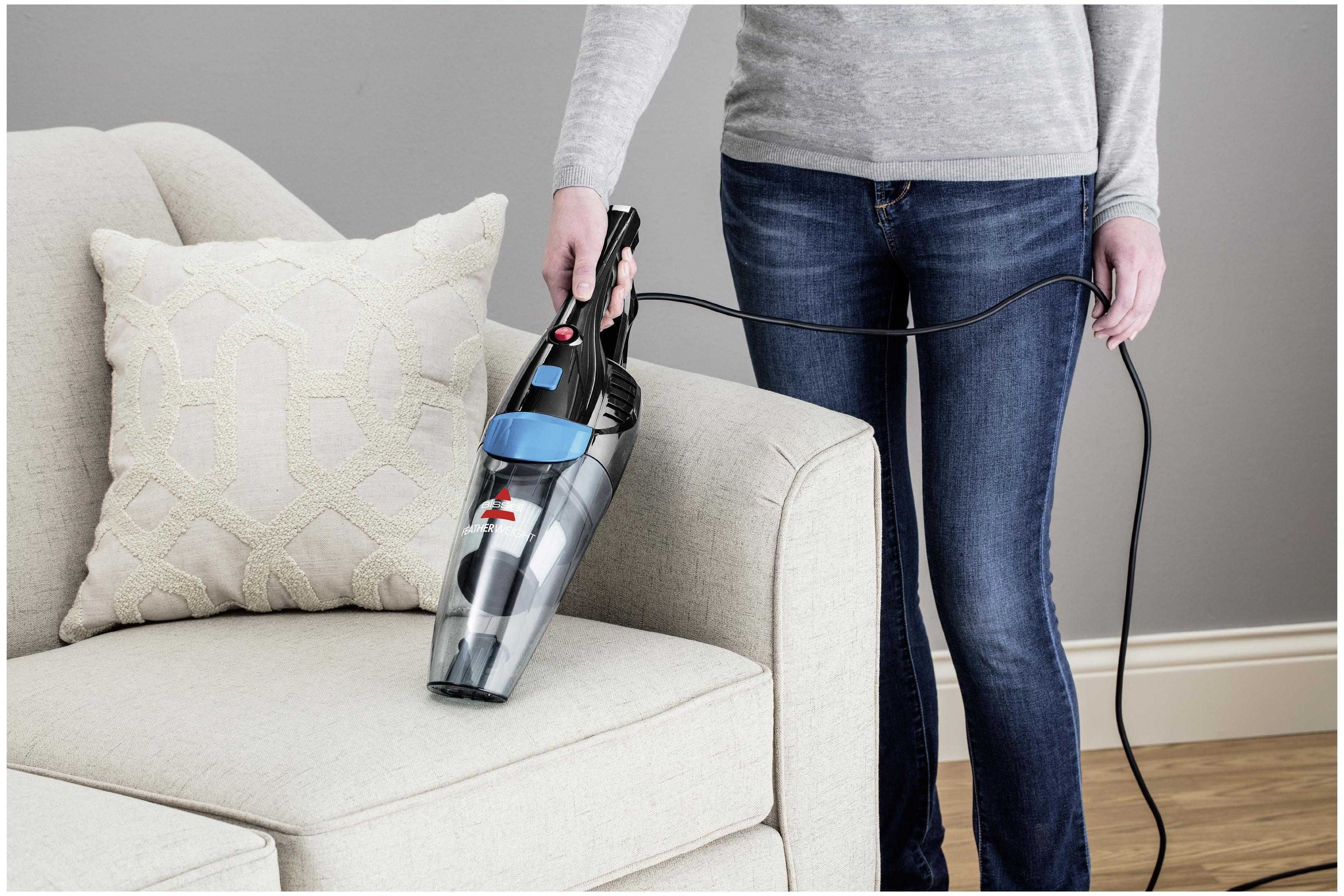 Eine Frau in Jeans saugt ein Sofa mit einem kleinen Handstaubsauger ab.