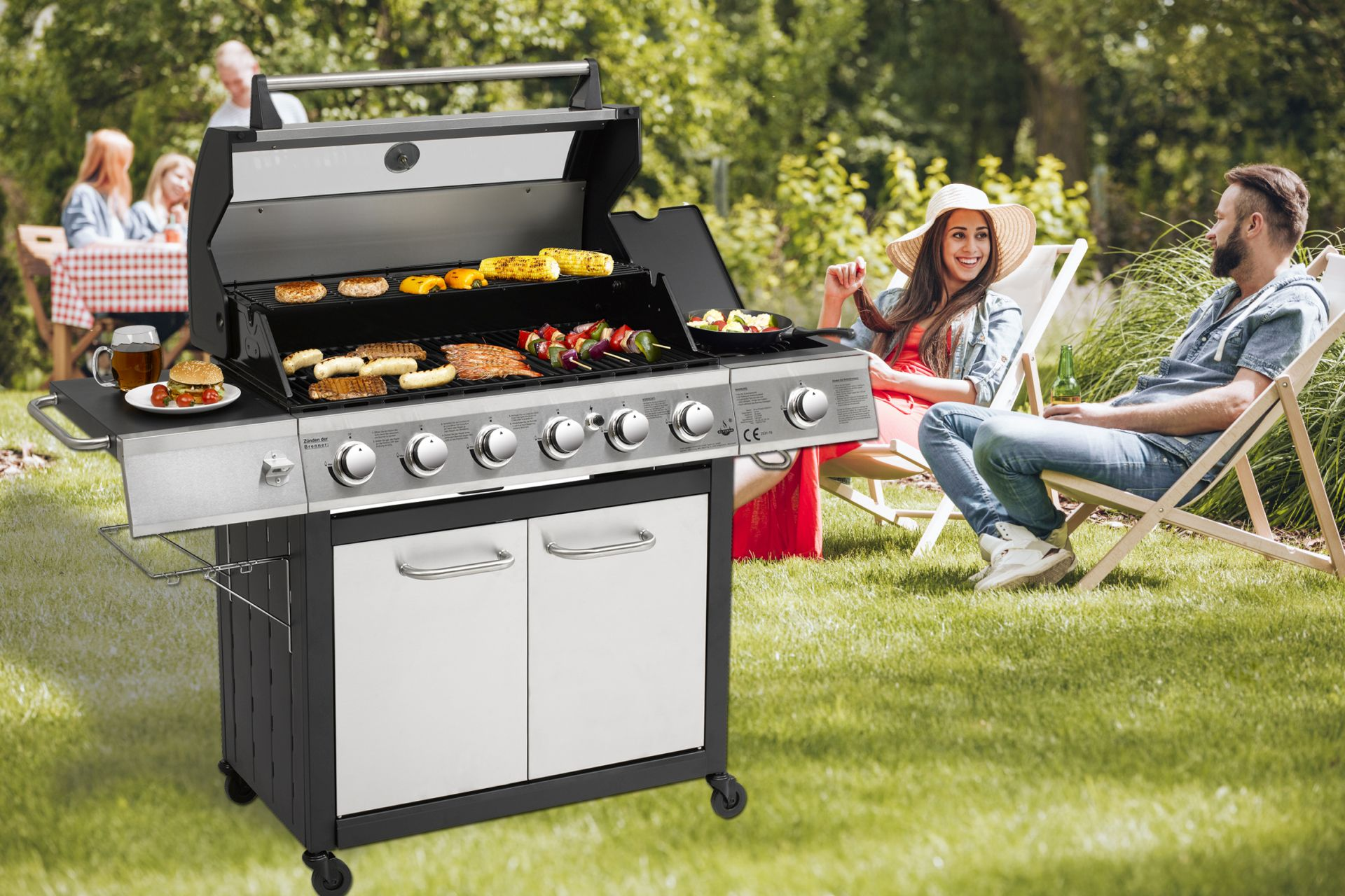 Ein Gasgrill mit darauf zubereitetem Essen. Ein Paar sitzt im Hintergrund an einem Tisch.