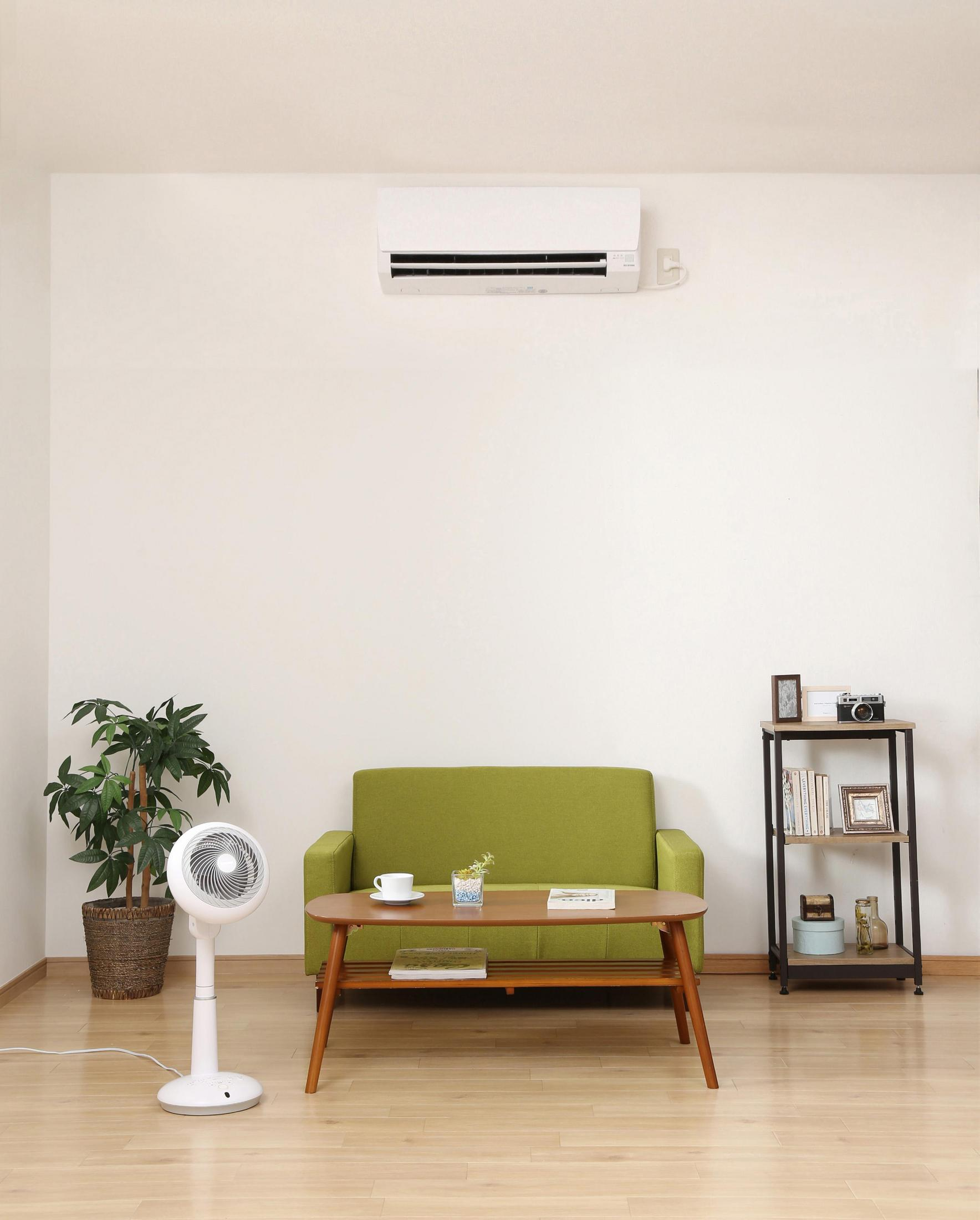 Salón con aire acondicionado en la pared sobre un sofá verde. También hay un ventilador y una mesa auxiliar.