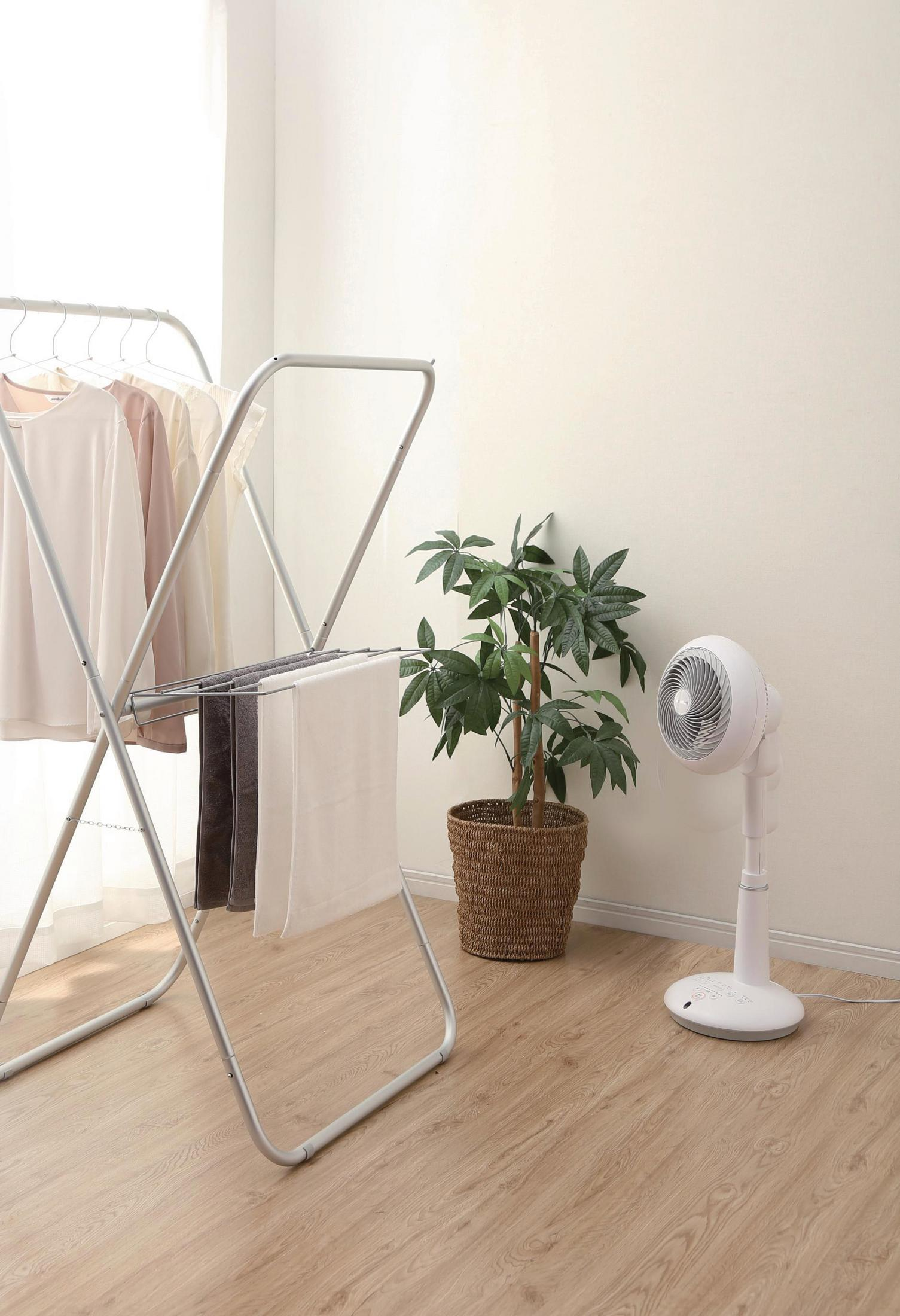 Un tendedero con ropa, una planta en maceta y un ventilador blanco en una habitación luminosa.