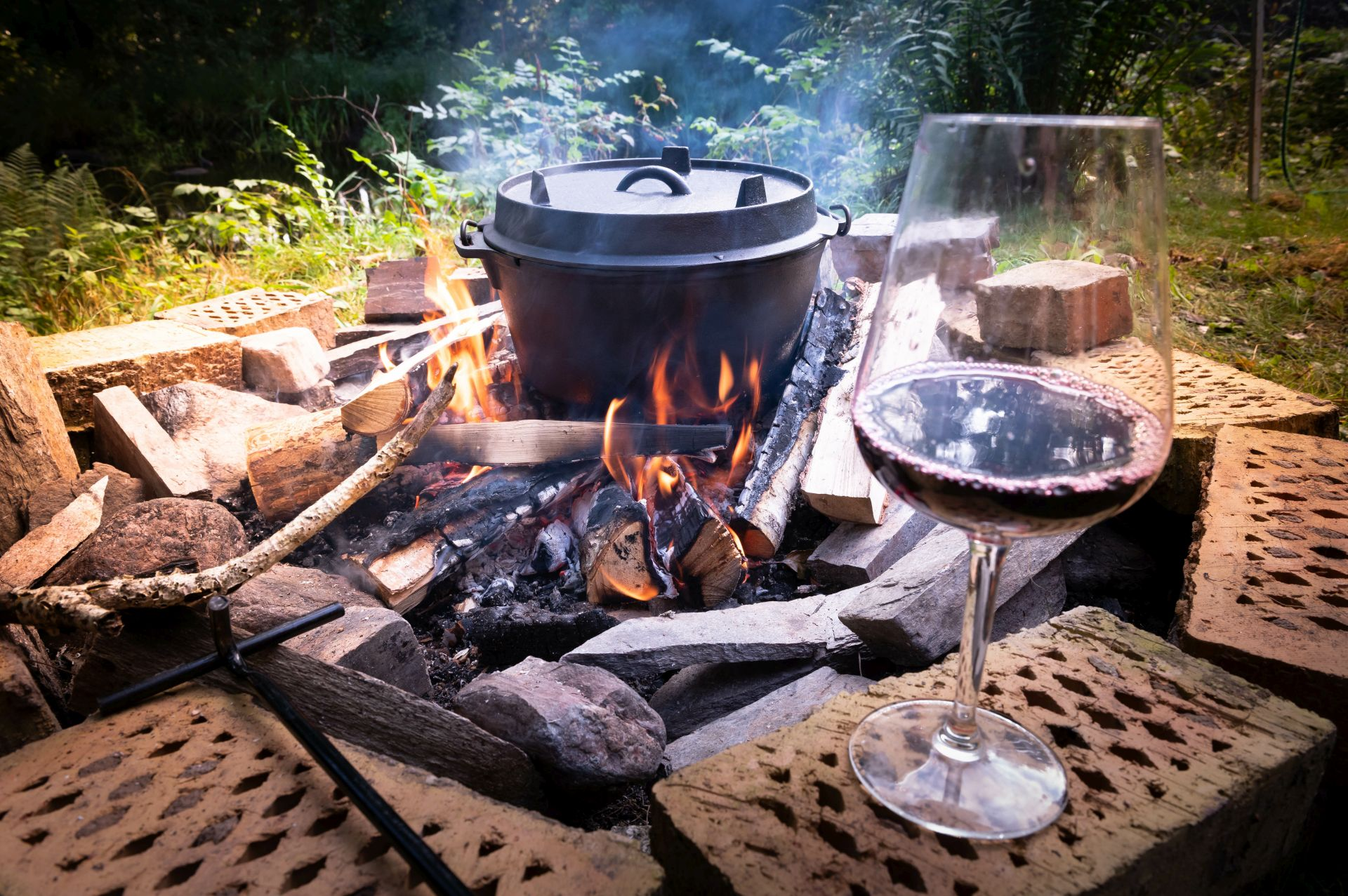 Lagerfeuer mit Topf und Rotweinglas, Feuer brennt, Wald im Hintergrund.