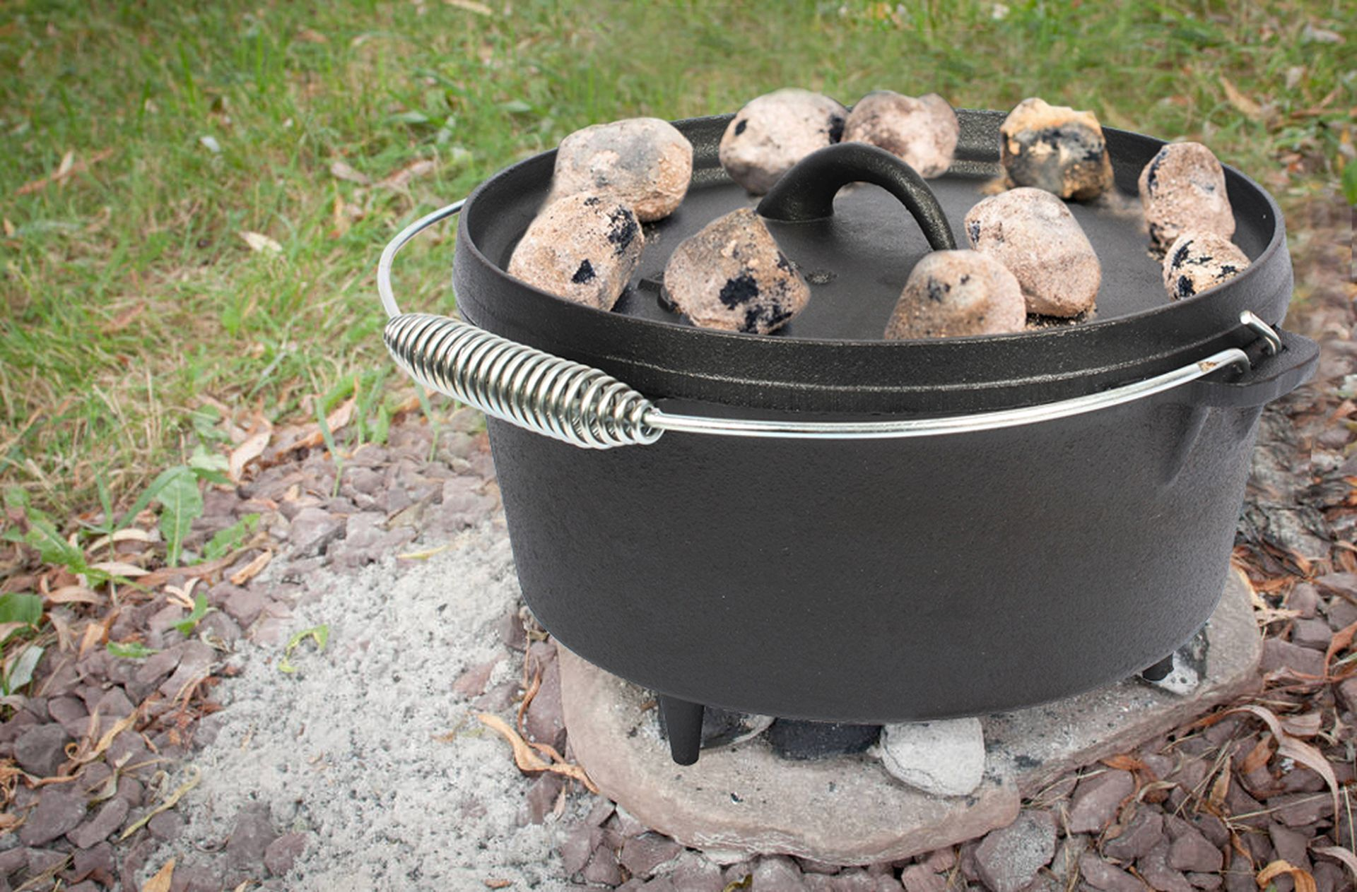 Dutch Oven mit Kohlen oben drauf. Es steht auf Steinen und Erde in der Nähe von Gras.