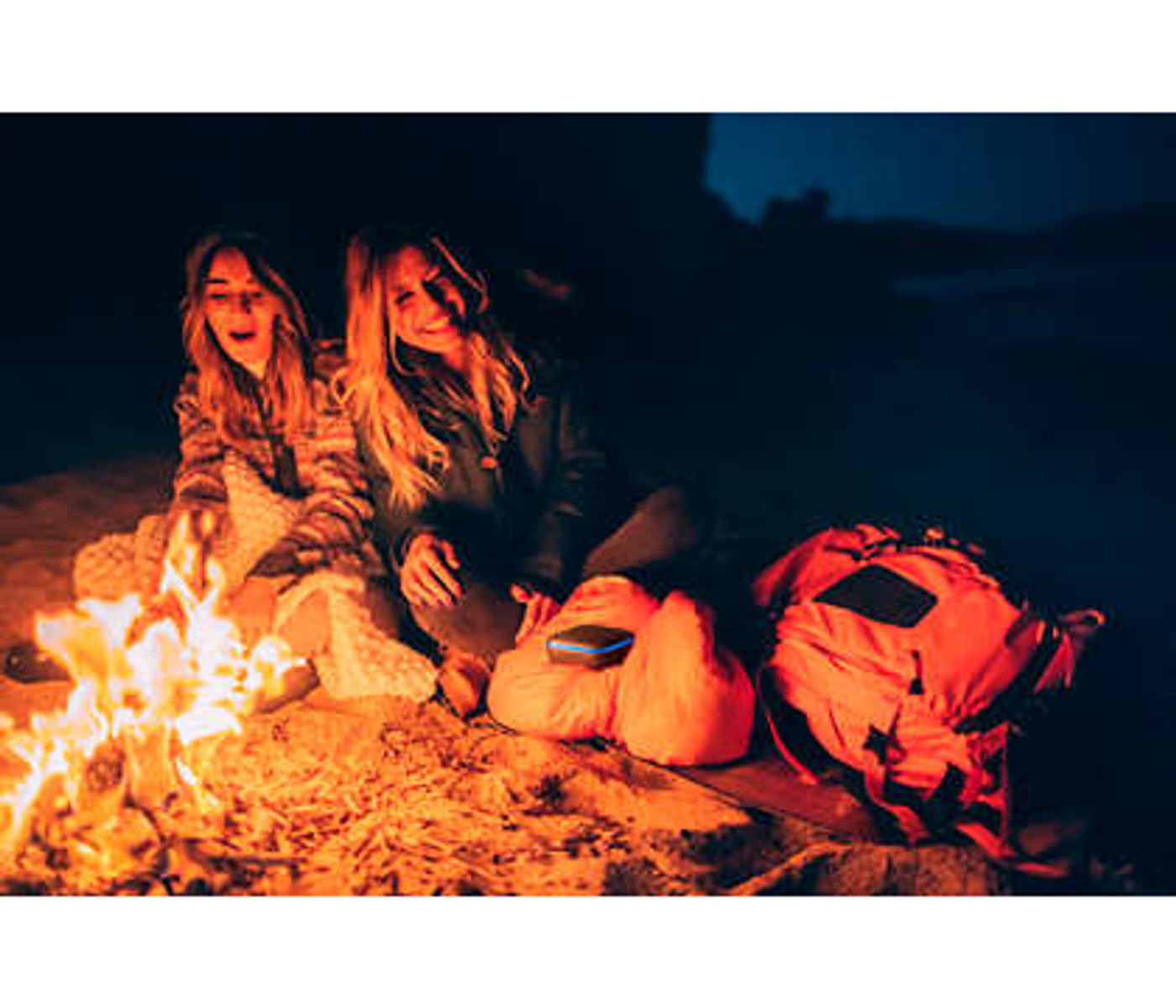 Dwie kobiety siedzą w nocy przy ognisku, z pomarańczowymi plecakami i głośnikiem.