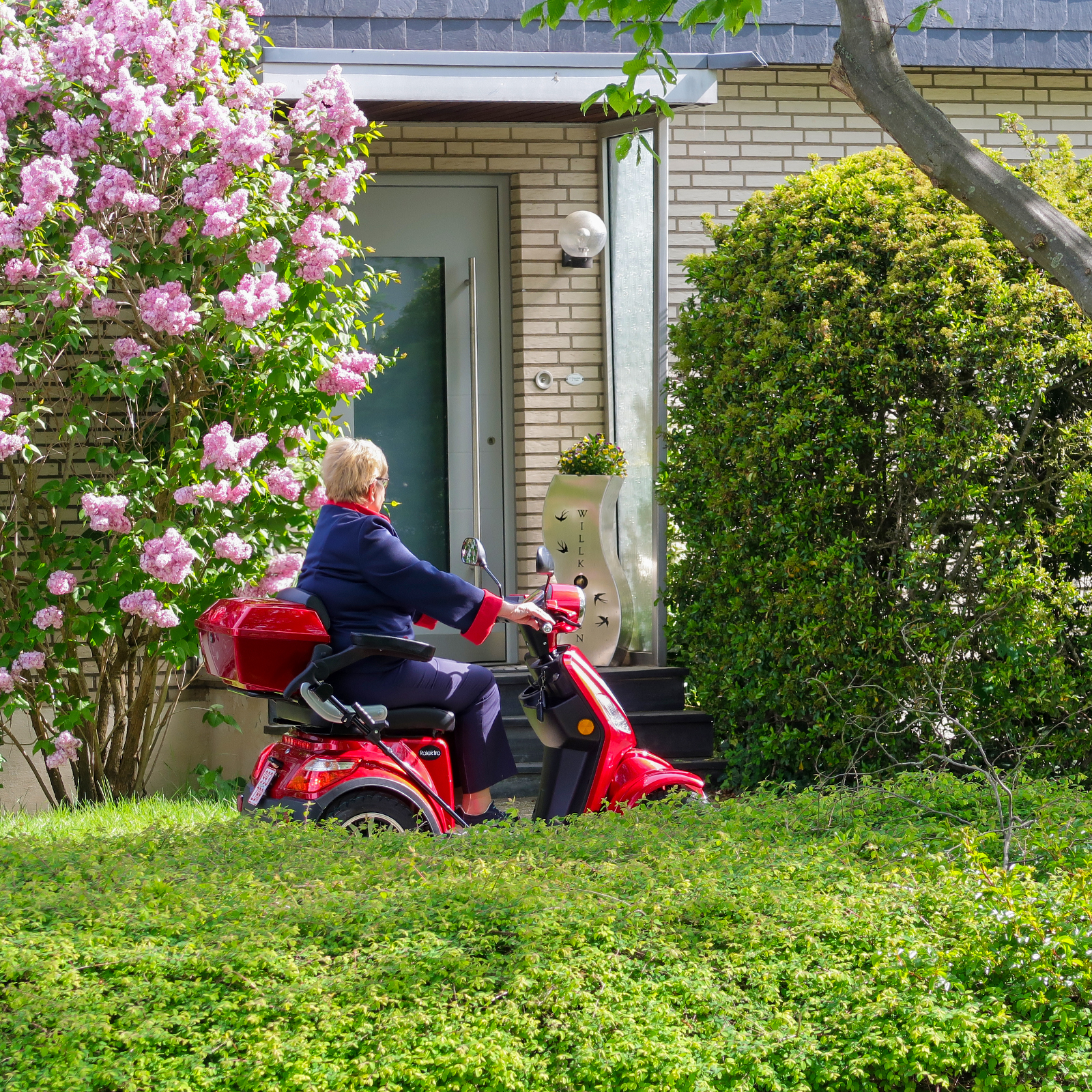 Frau auf rotem Roller an Haus mit blühenden Bäumen und Büschen. Grünes Gras.