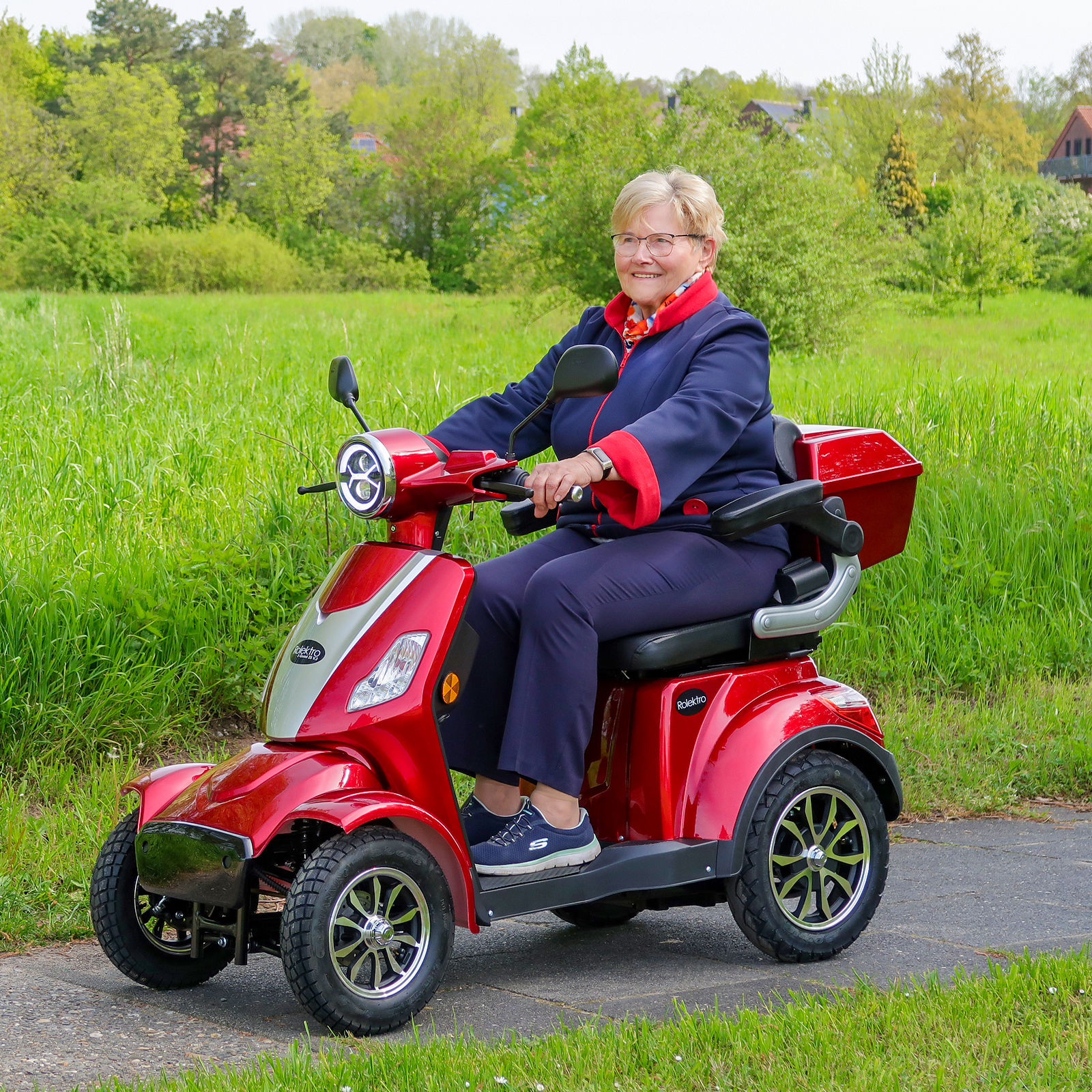 Eine ältere Frau auf einem roten Mobilitätsroller fährt auf einem Weg durch grünes Gras.