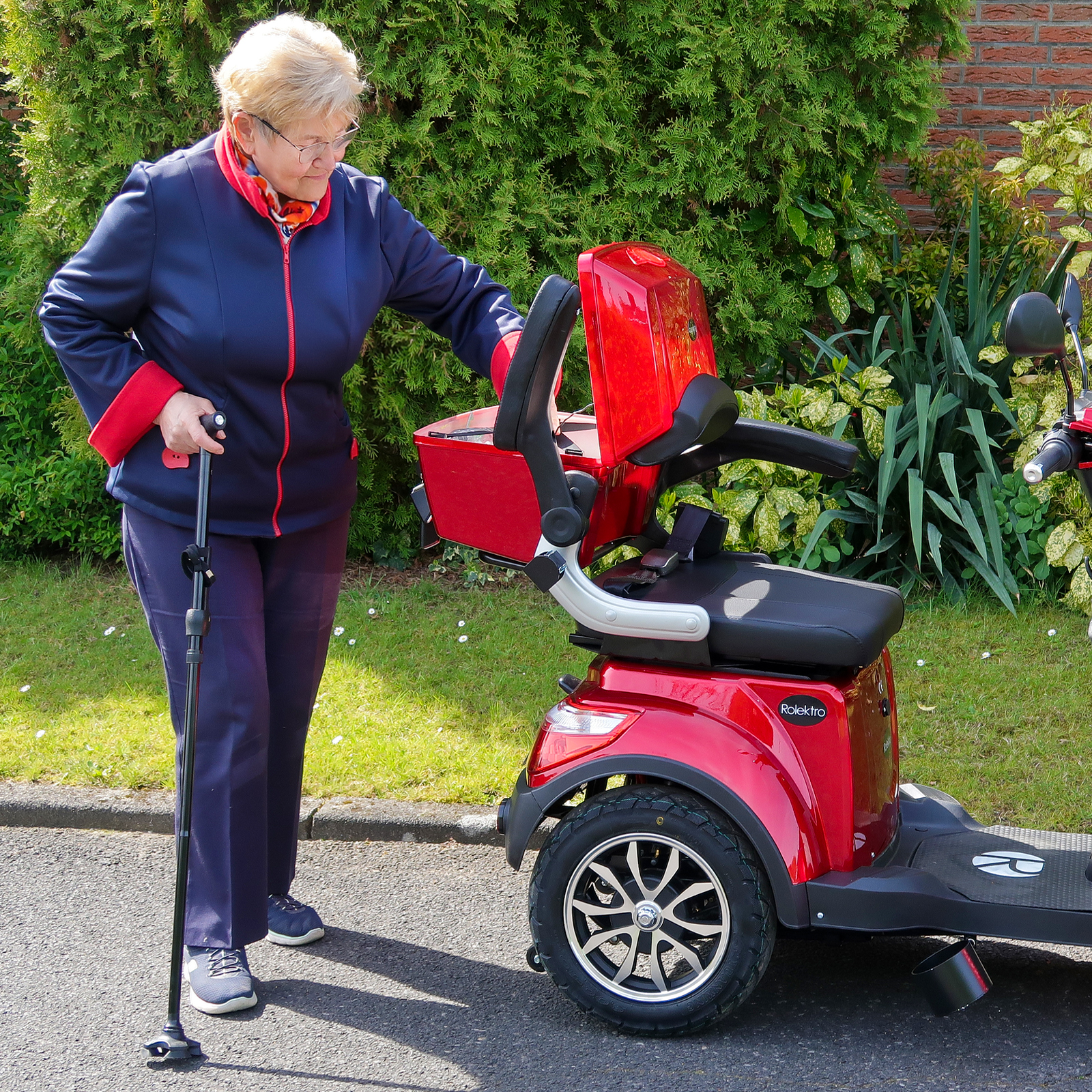 Ältere Frau mit Gehstock in der Nähe eines roten Mobilitätsrollers. Sie trägt eine blaue Jacke.