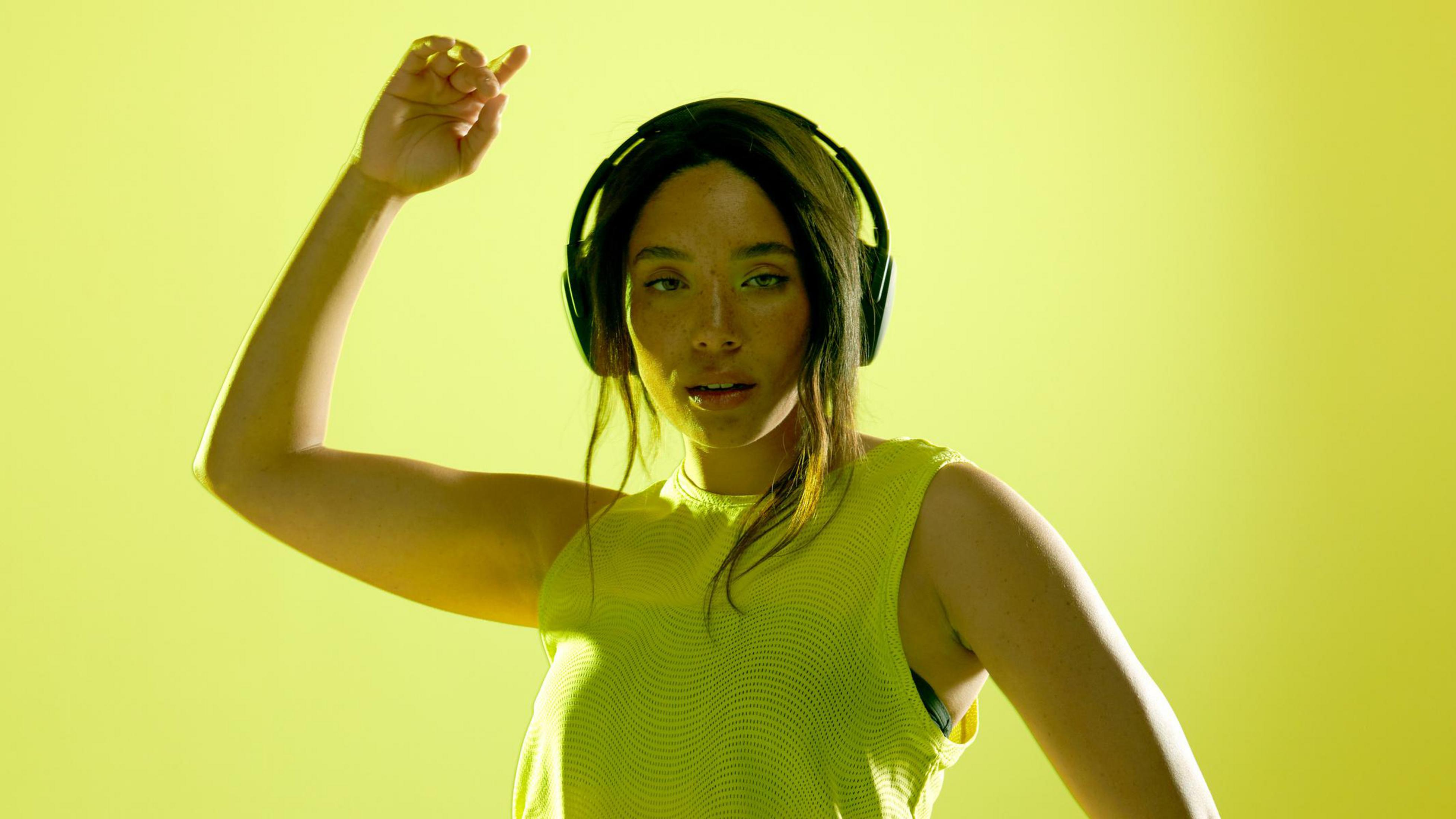 Kobieta w żółtym topie i słuchawkach, uniesiona ręka. Żółte tło.