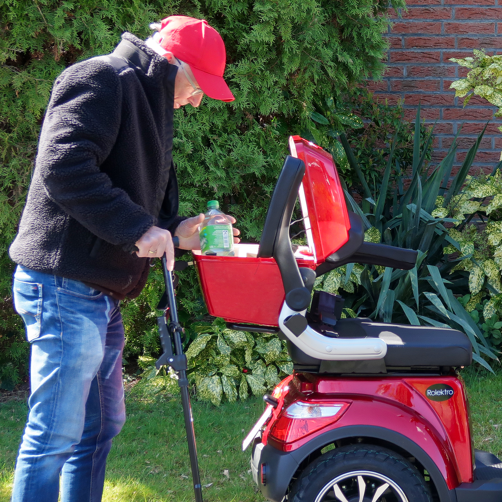 Ein älterer Mann mit roter Mütze neben einem roten Mobilitätsroller, hält einen Stock und eine Wasserflasche.