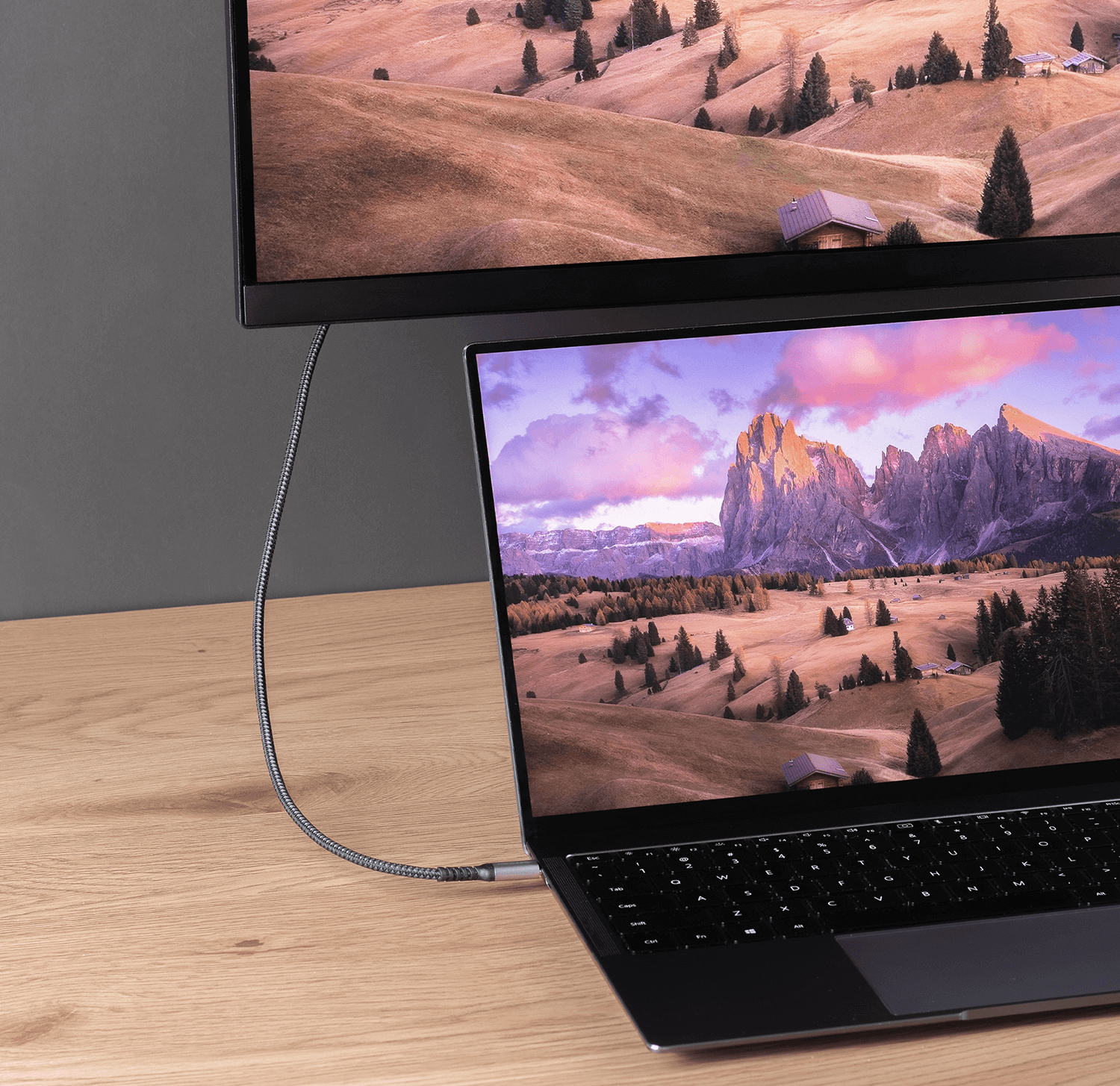 Laptop podłączony do monitora za pomocą kabla. Monitor wyświetla krajobraz górski. Laptop jest na drewnie.