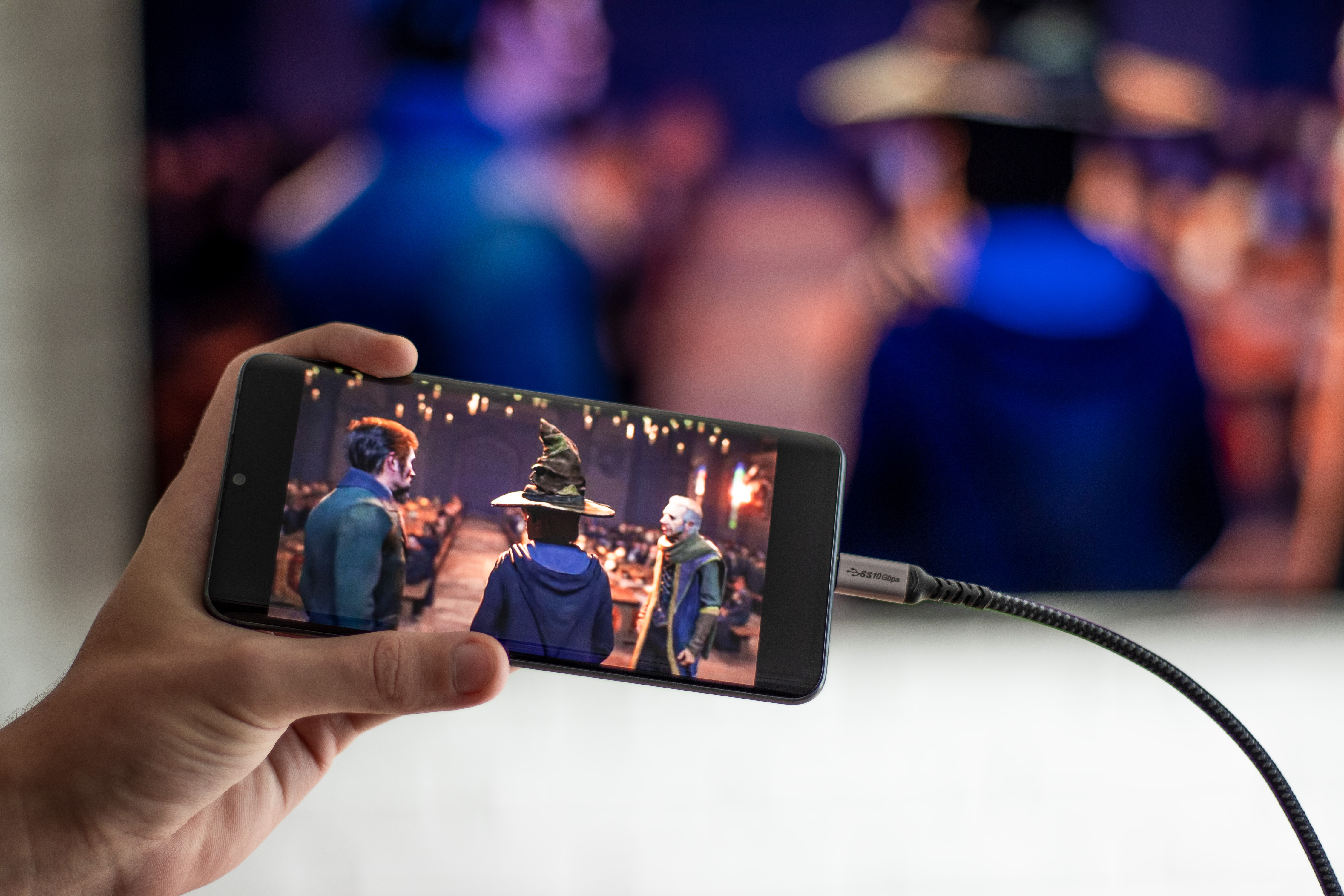 Ręka trzymająca telefon wyświetlający scenę filmową. Telefon jest podłączony do większego ekranu pokazującego ten sam obraz.