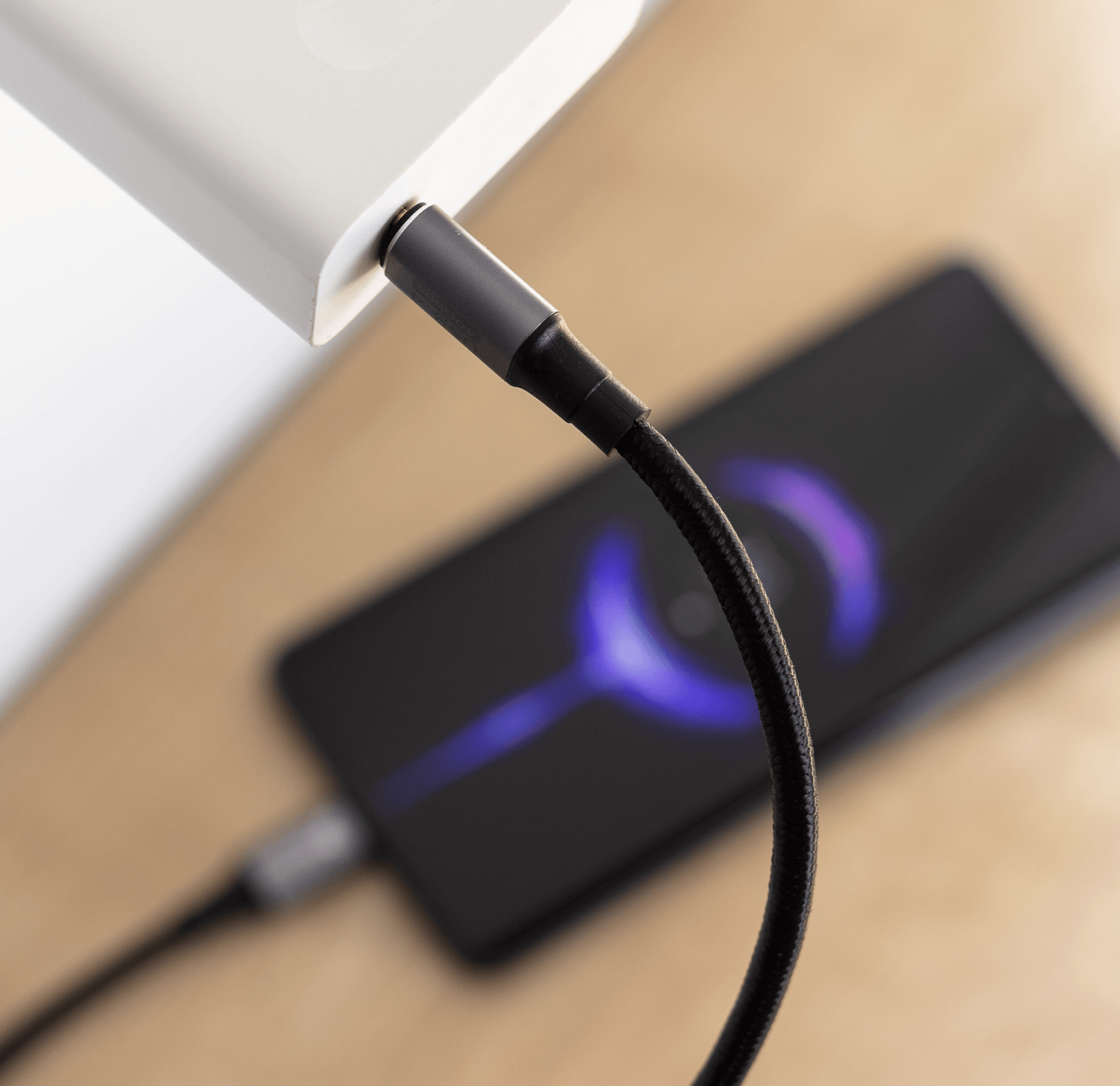 Telefon ładuje się czarnym kablem. Podłączona ładowarka. Drewniana powierzchnia.