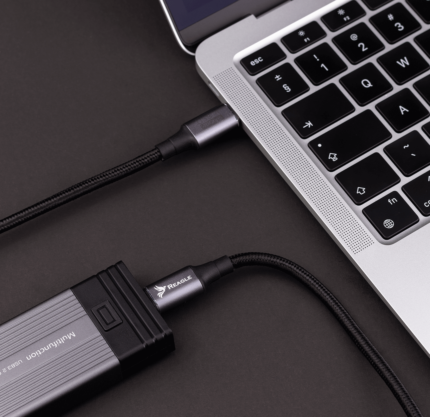 Laptop podłączony do srebrnego kabla USB-C. Czarny kabel podłączony do czarno-szarego urządzenia. Czarne tło.