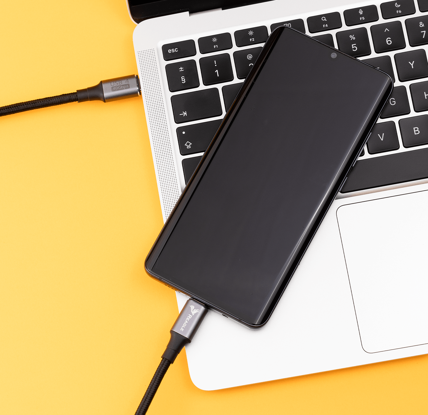 Laptop z czarnym smartfonem podłączonym kablem USB-C na żółtej powierzchni.
