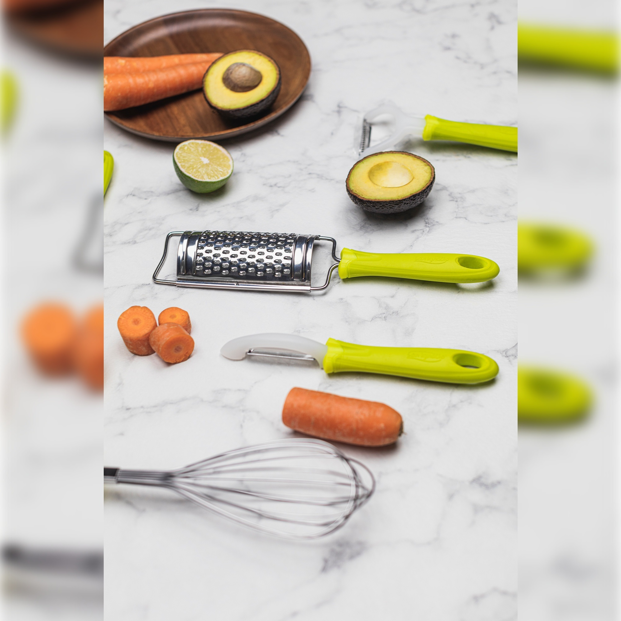 Utensilios de cocina: zanahorias, aguacate, rallador, pelador, batidor sobre superficie de mármol.