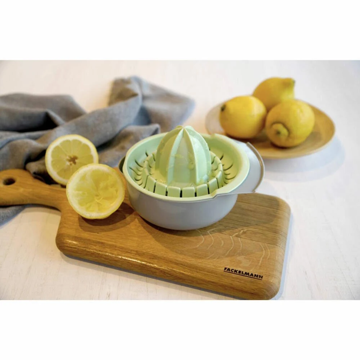 Scena cucina con spremiagrumi, limoni, tagliere e panno grigio su superficie bianca.