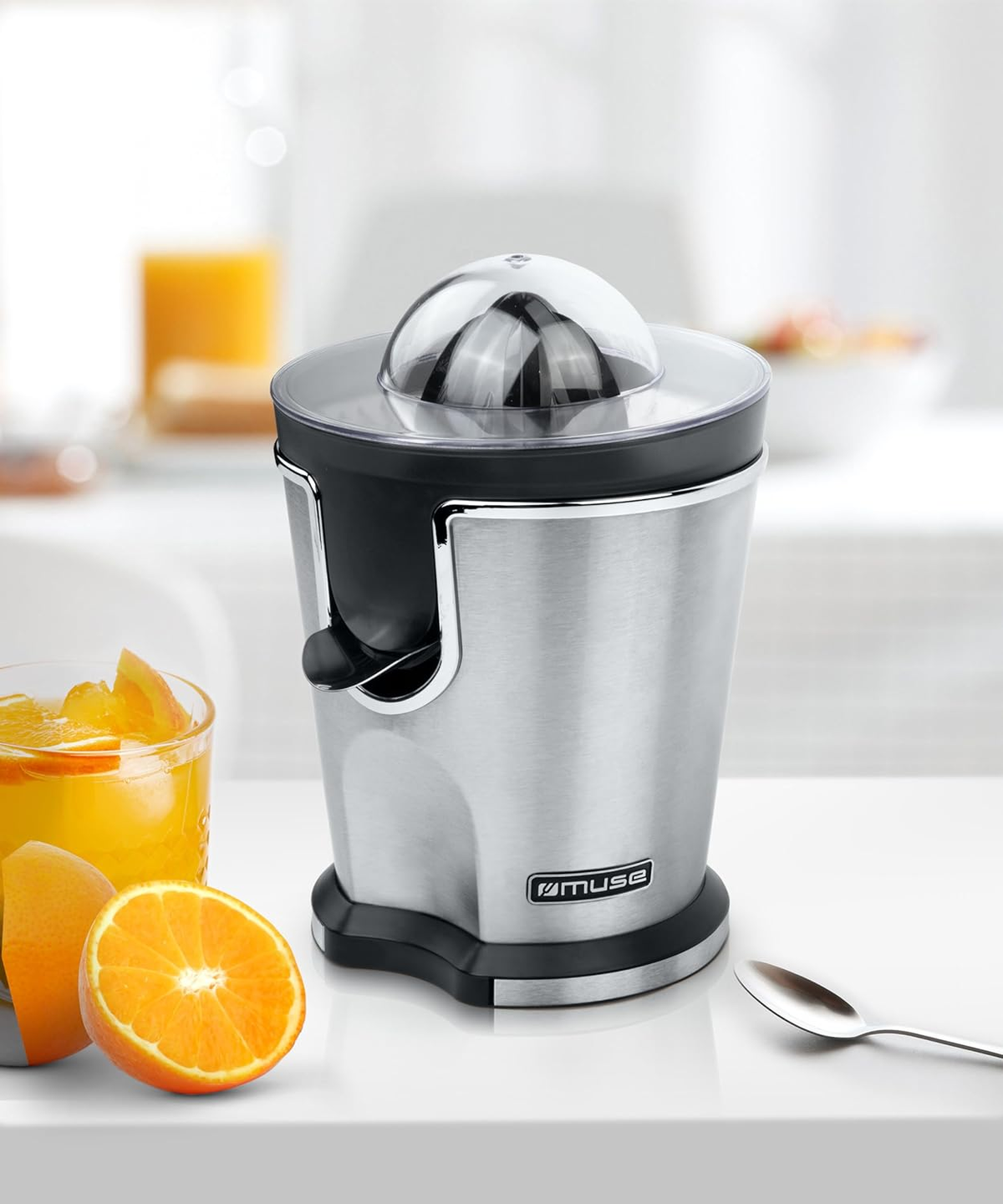 Una exprimidora de cítricos Muse plateada con naranjas y un vaso de zumo. Una cuchara en la mesa.