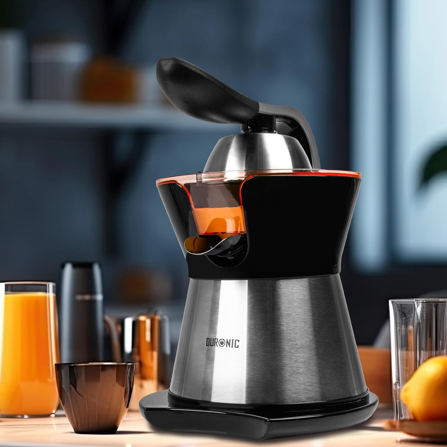 Exprimidor con diseño negro y plateado, jugo de naranja dentro. Otros vasos visibles.