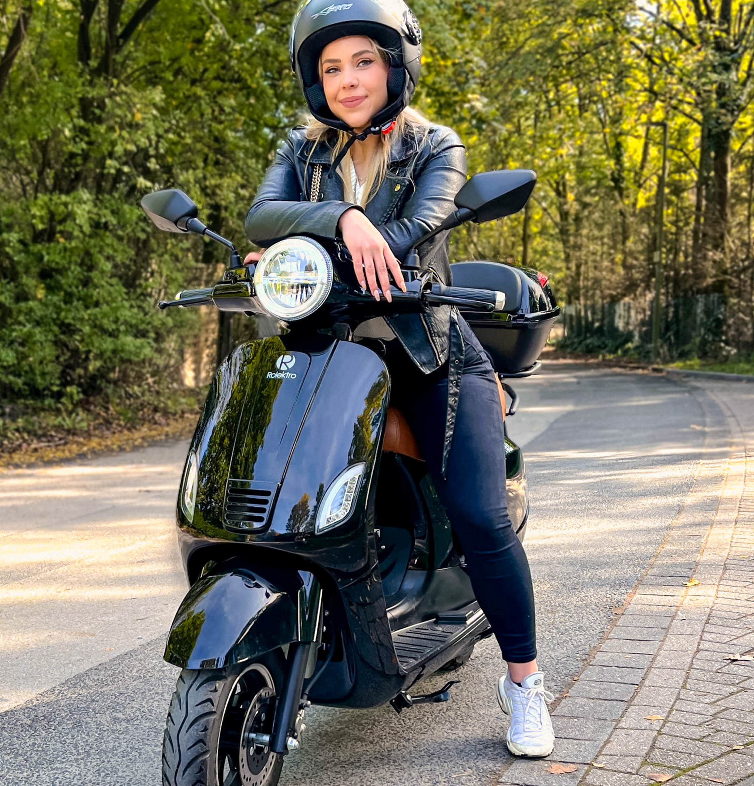 Eine Frau auf einem schwarzen Roller mit Helm und Lederjacke auf einer Straße mit Bäumen.