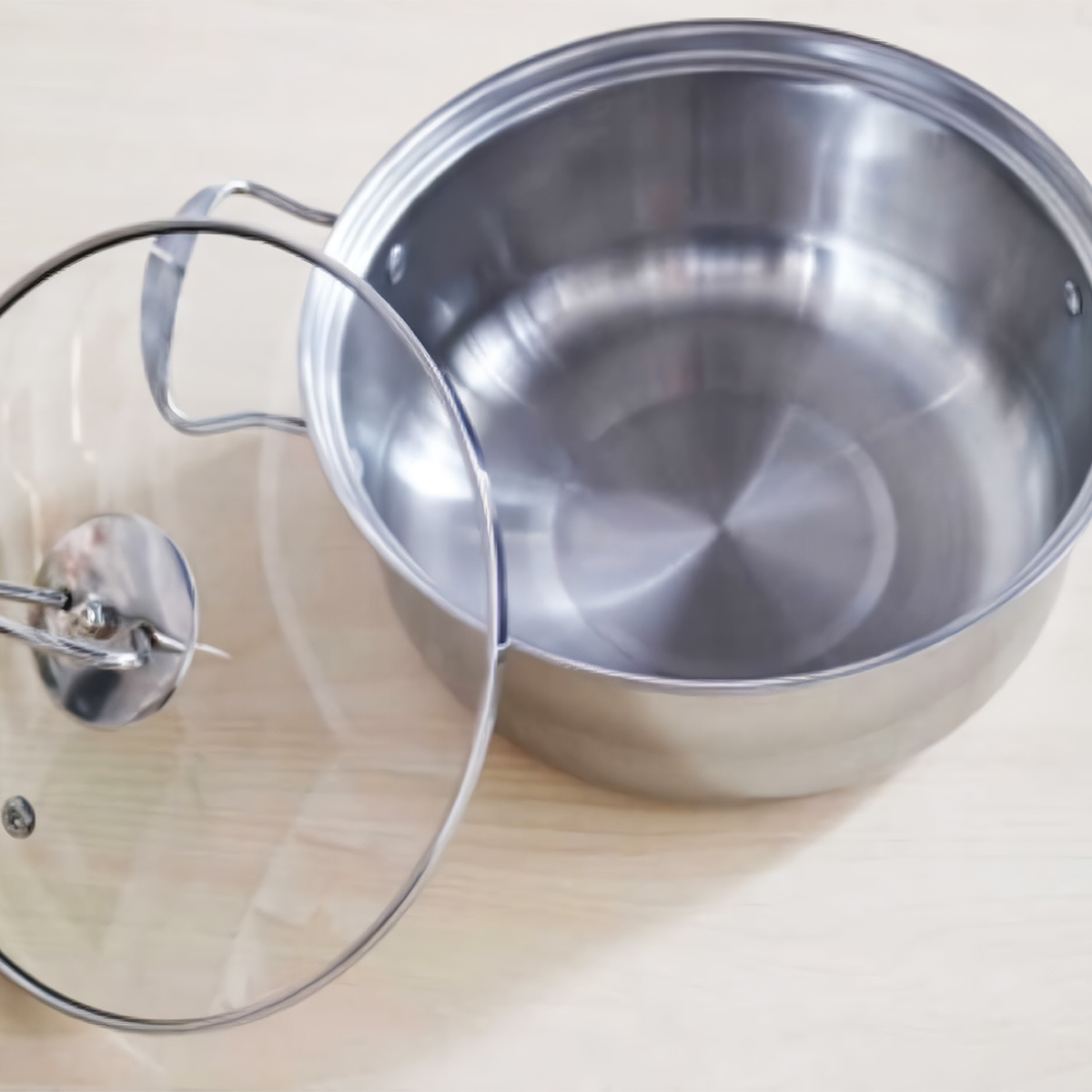 Olla de acero inoxidable con tapa de cristal sobre una superficie de madera clara. Vacia.