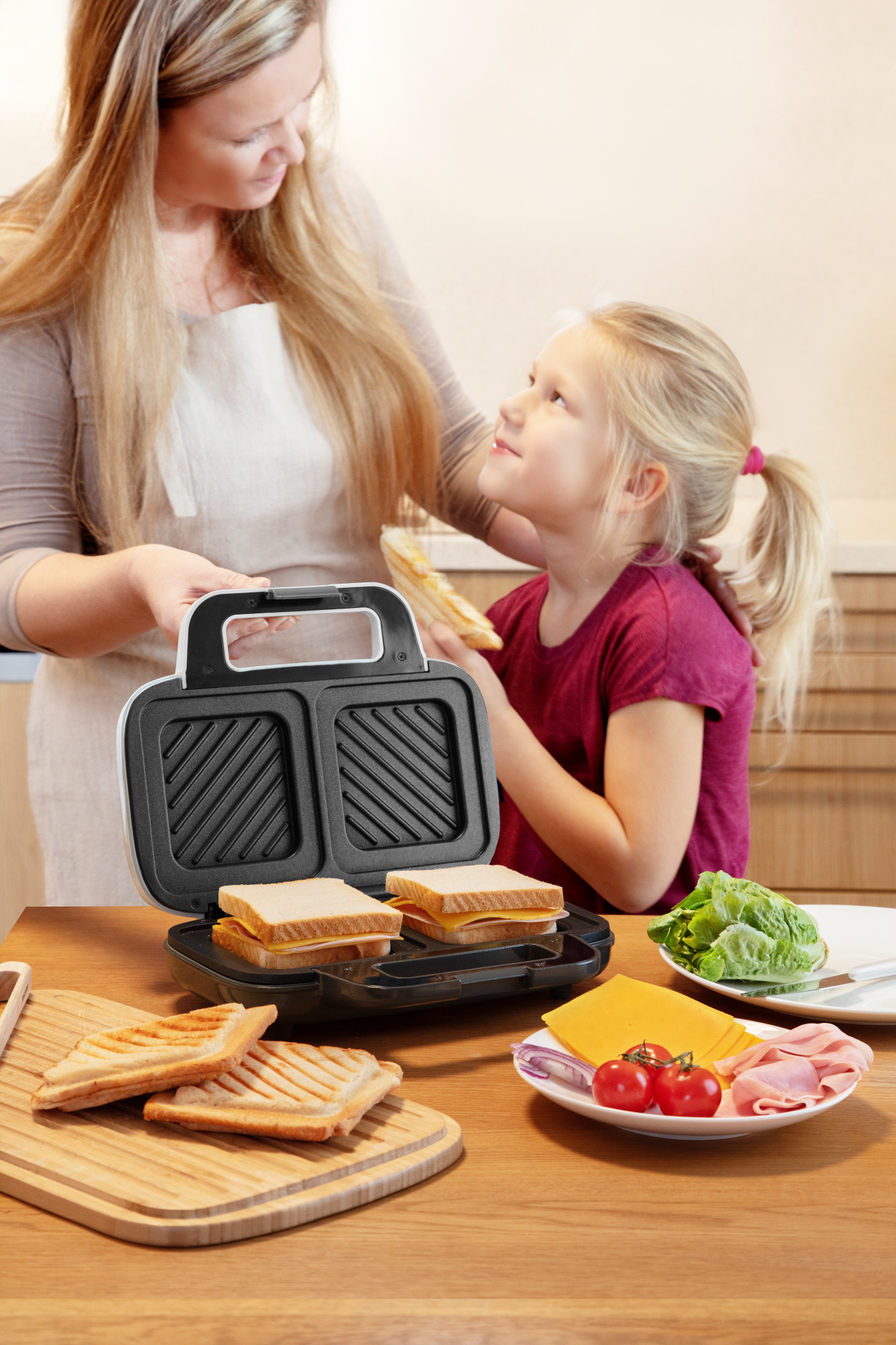 Frau und Mädchen machen Sandwiches mit einem Sandwichmaker. Zutaten auf dem Tisch.
