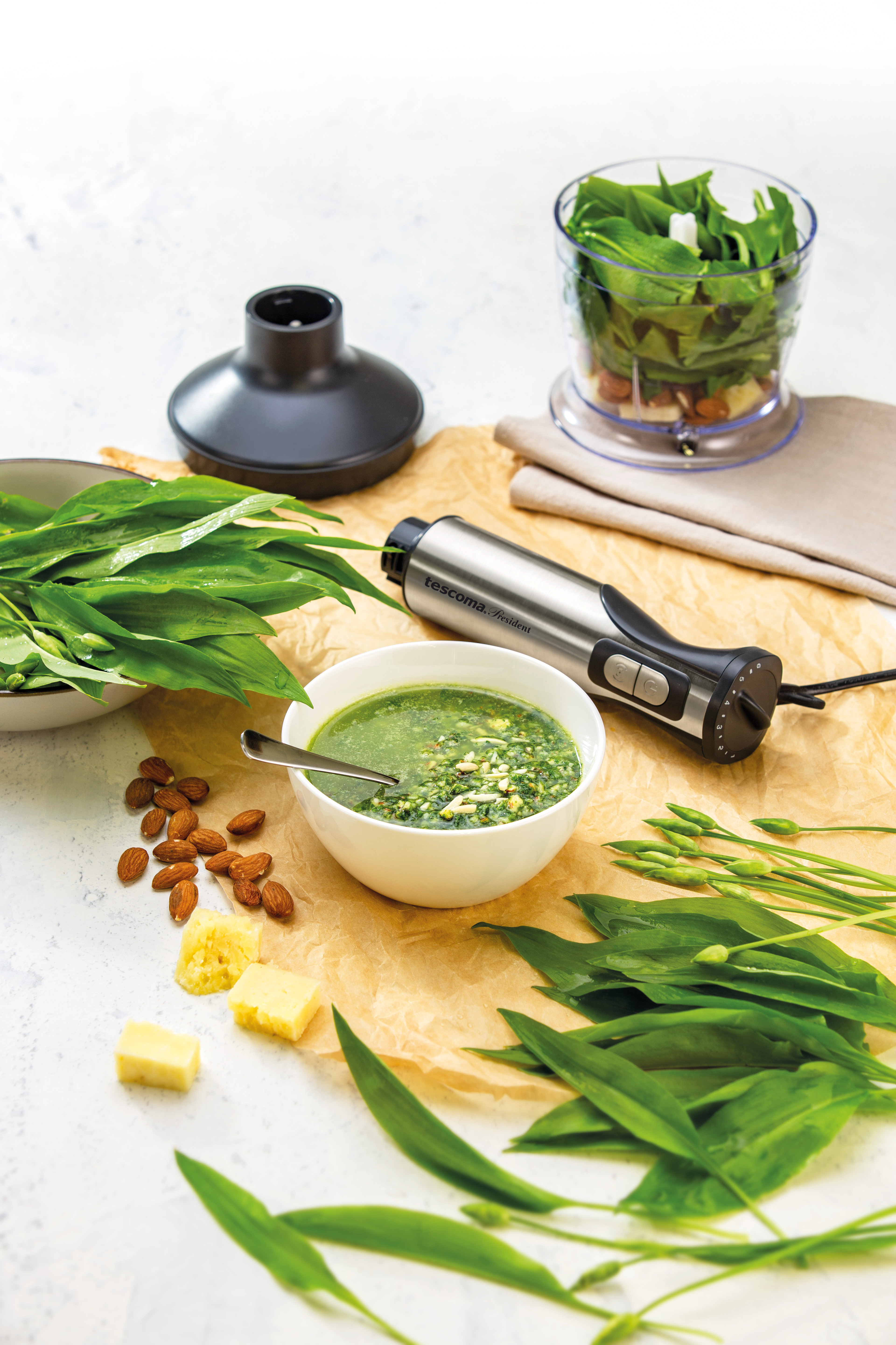 Mixer mit Pesto in einer Schüssel, umgeben von Zutaten wie Basilikum und Mandeln.
