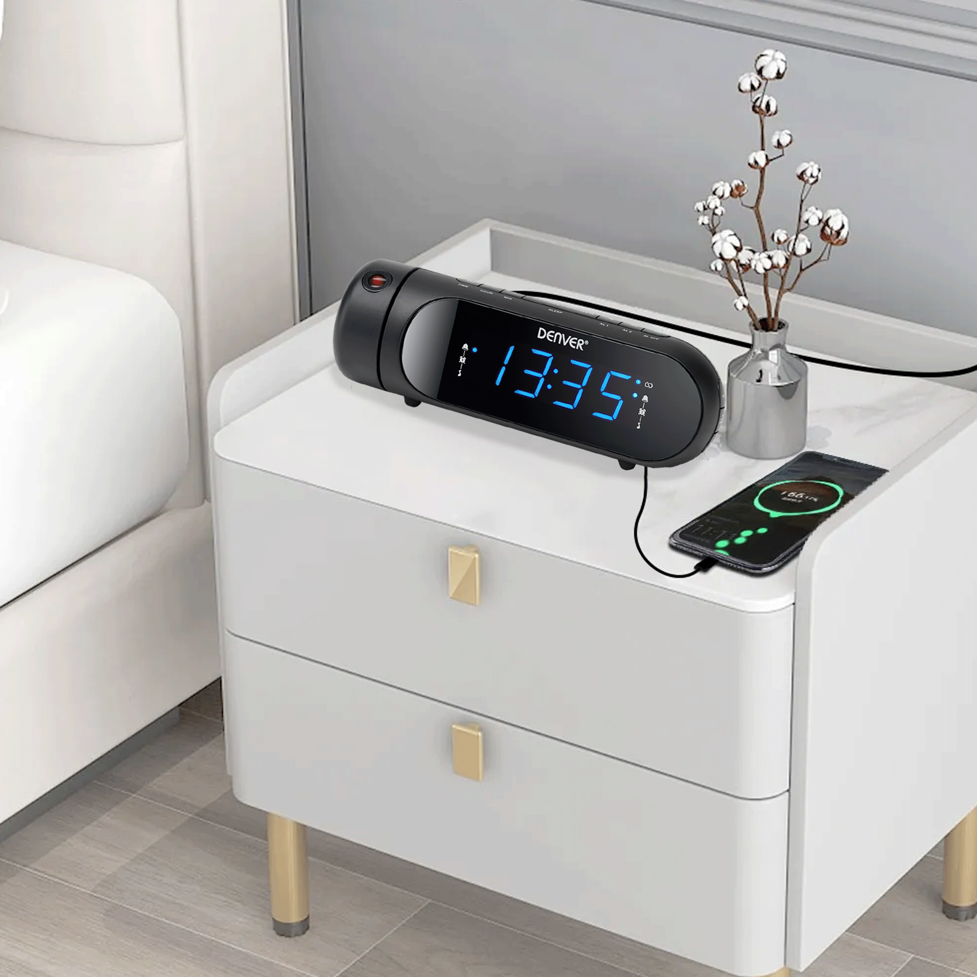 Czarny budzik z niebieskim wyświetlaczem na białej szafce nocnej. Telefon obok się ładuje.
