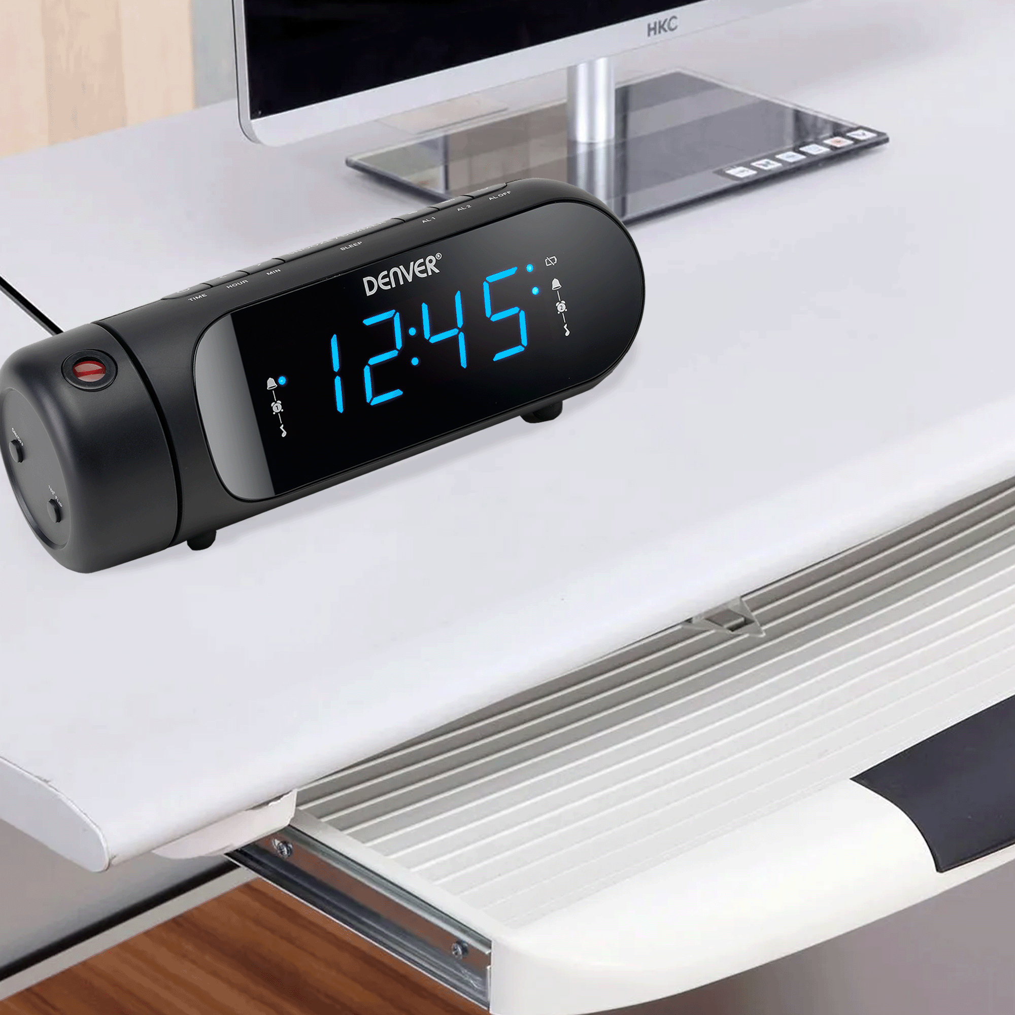 Czarny cyfrowy budzik na białym biurku. Pokazuje godzinę 12:45 w niebieskim świetle.