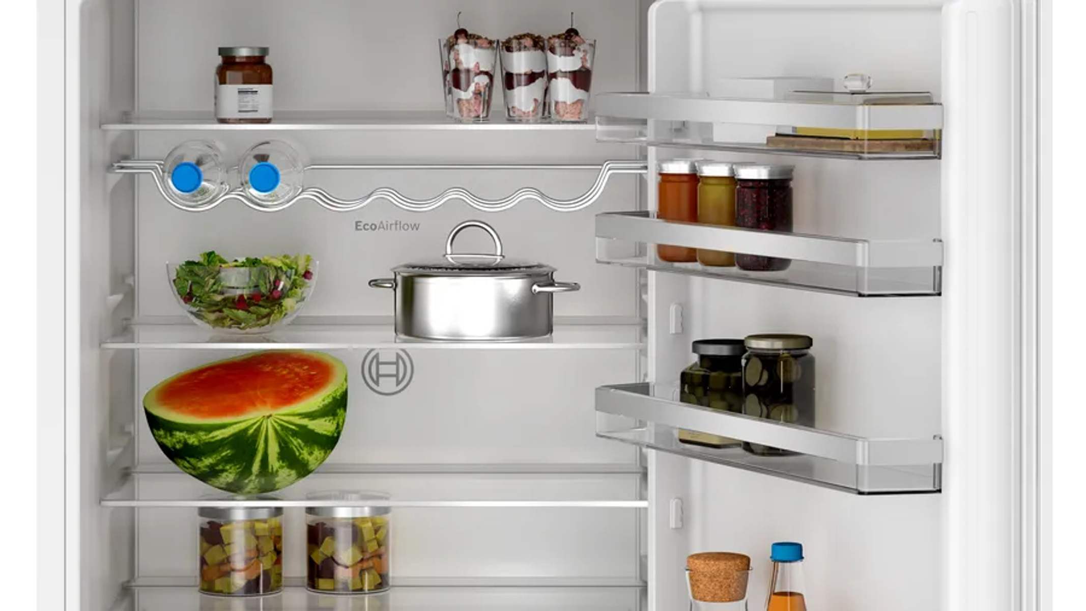 Dentro de un refrigerador con una sandía, frascos, una olla y ensalada fresca.