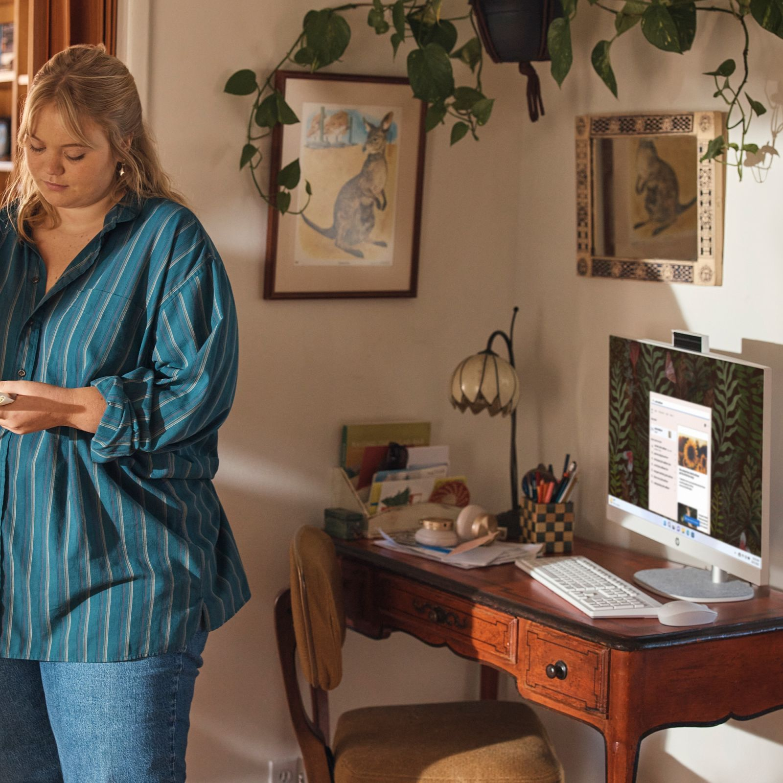 Kobieta w koszuli w paski stoi obok biurka z komputerem, zdjęcia na ścianie.