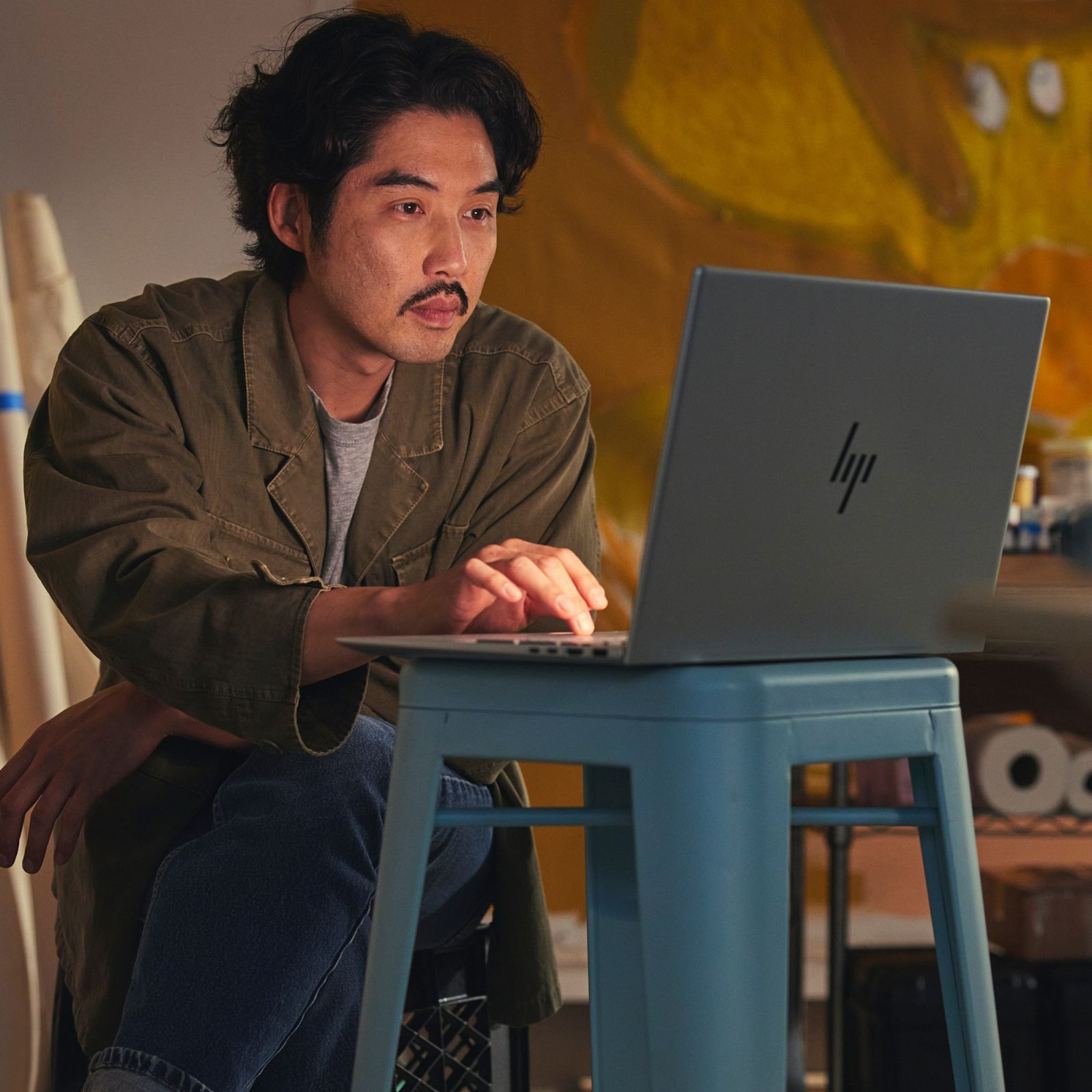 Mężczyzna z wąsami używa laptopa na niebieskim stołku. Tło: grafika.