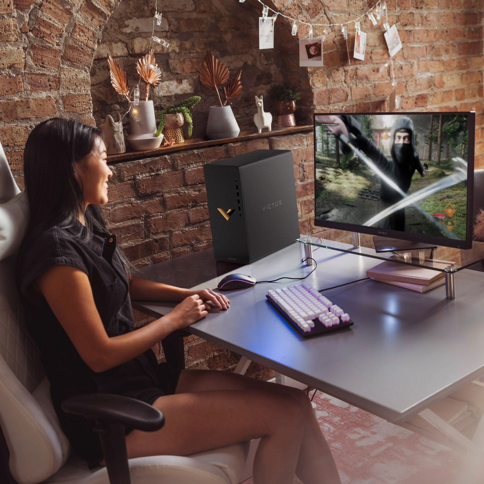 Kobieta gra przy biurku z czarnym komputerem, monitorem, klawiaturą i myszką. Tło z ceglanej ściany.
