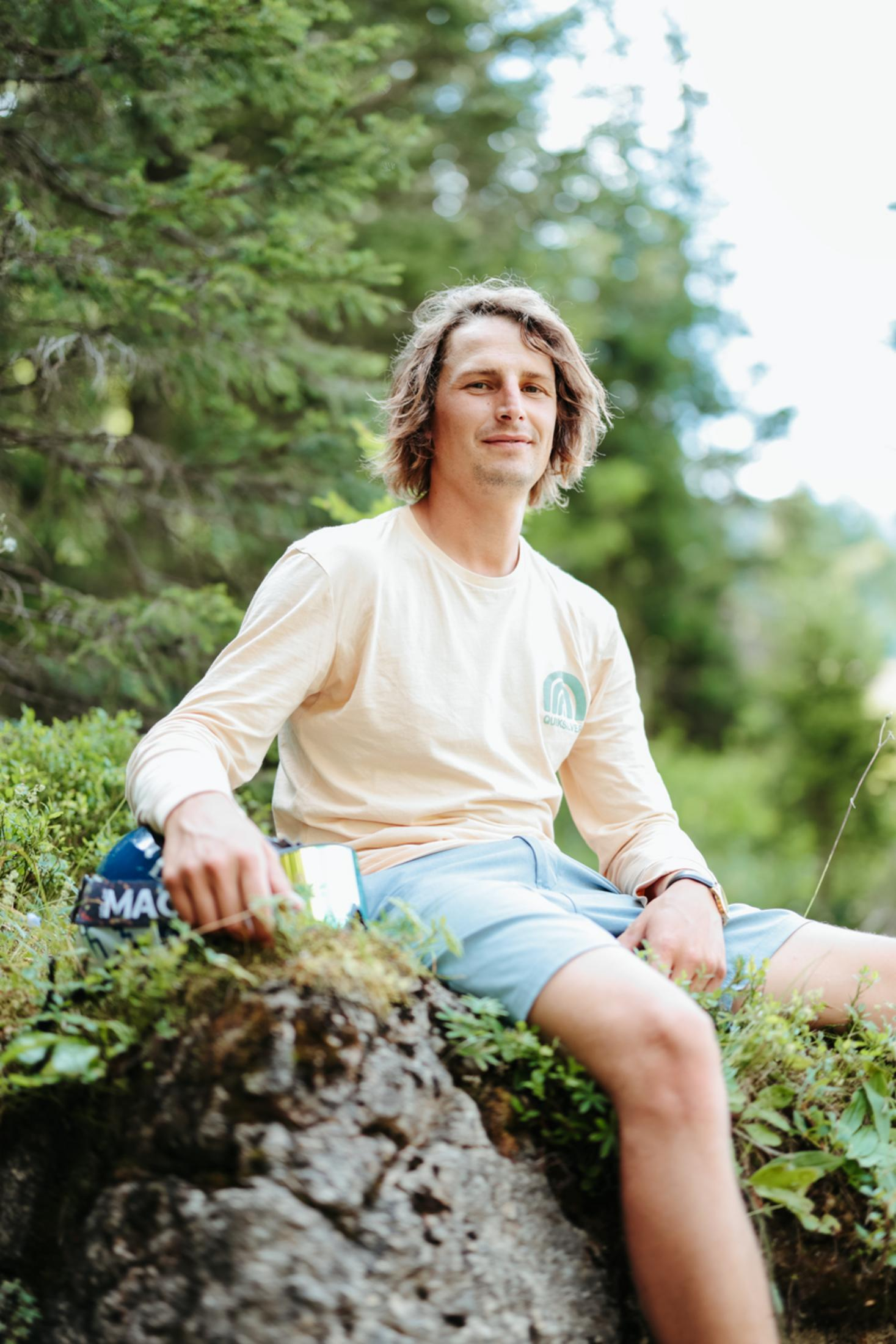 A man sits on a rock in nature, wearing a long-sleeved shirt.