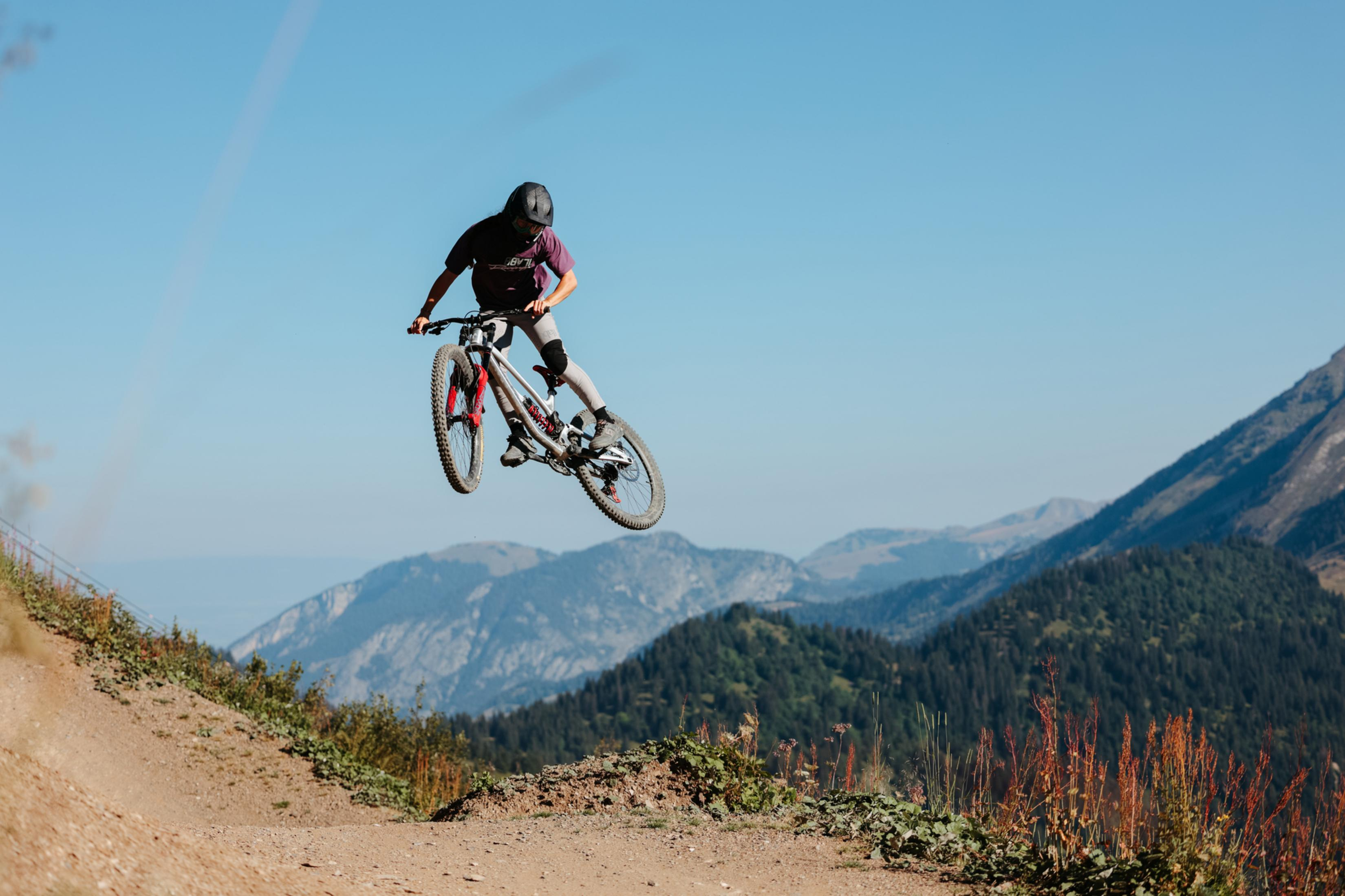 Mountain biker in mid-air, performing a jump. Blue sky, green mountains in the background.