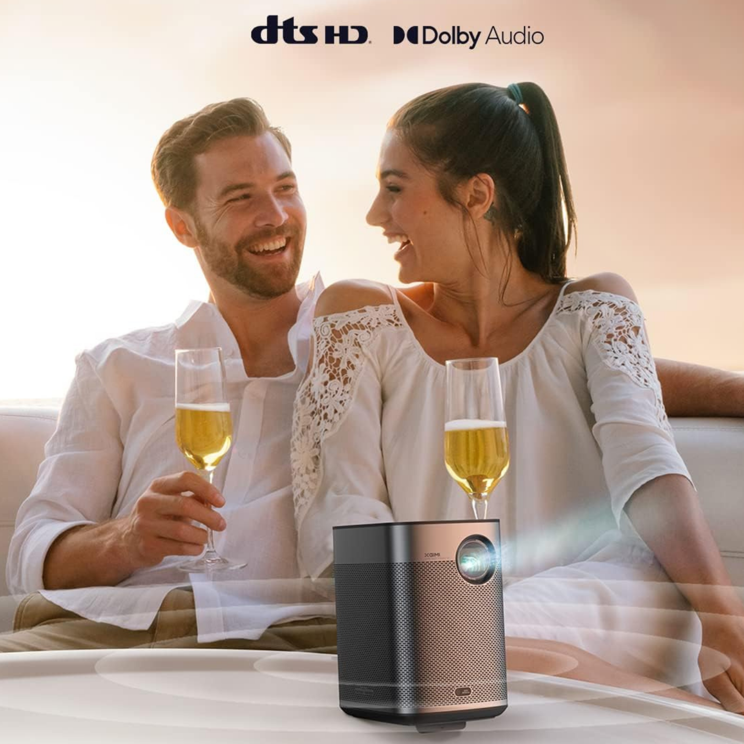 A couple smiles while on a boat, with a black and gold projector and two glasses of champagne.