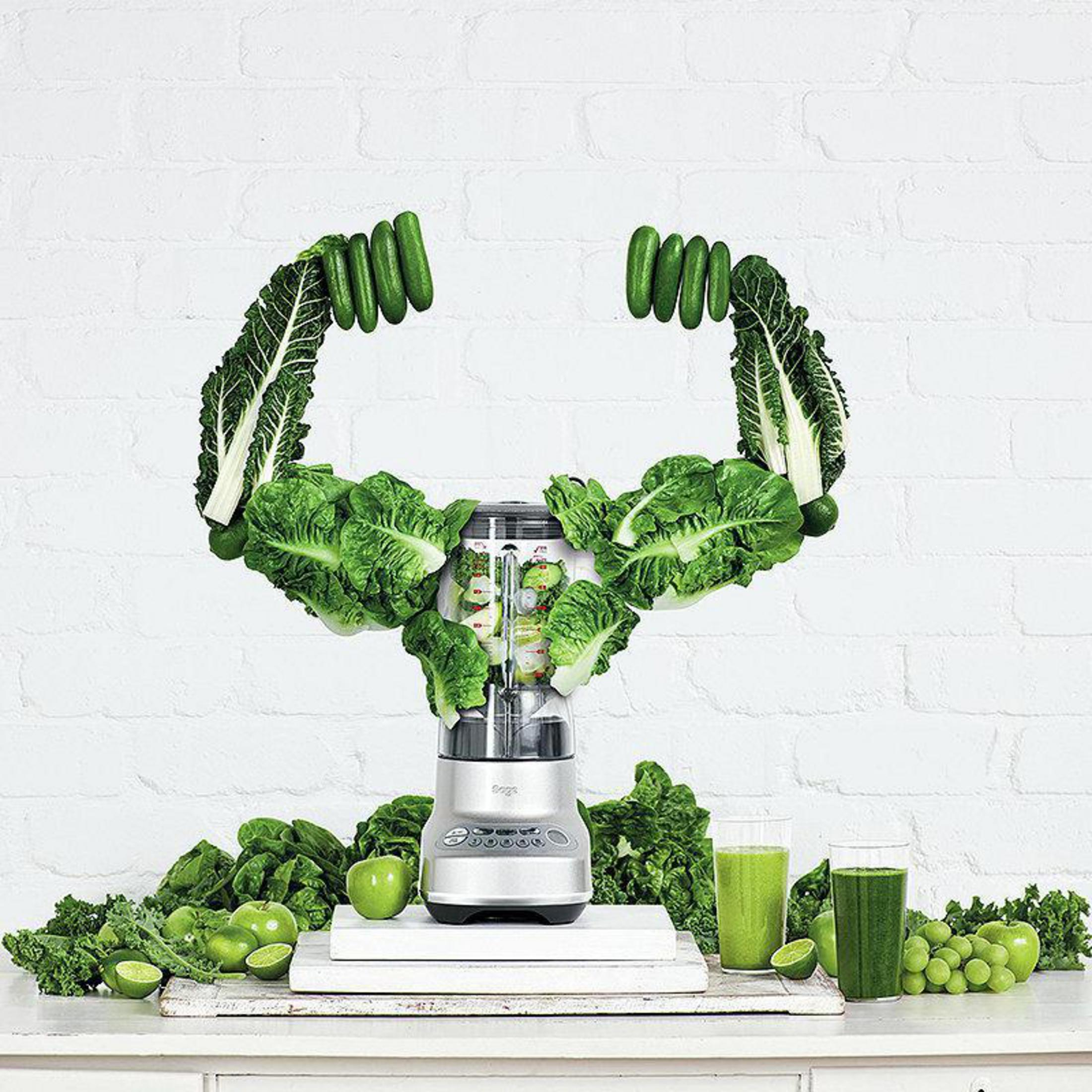 A blender with green vegetables arranged in the shape of arms. Two glasses of juice are on the table.