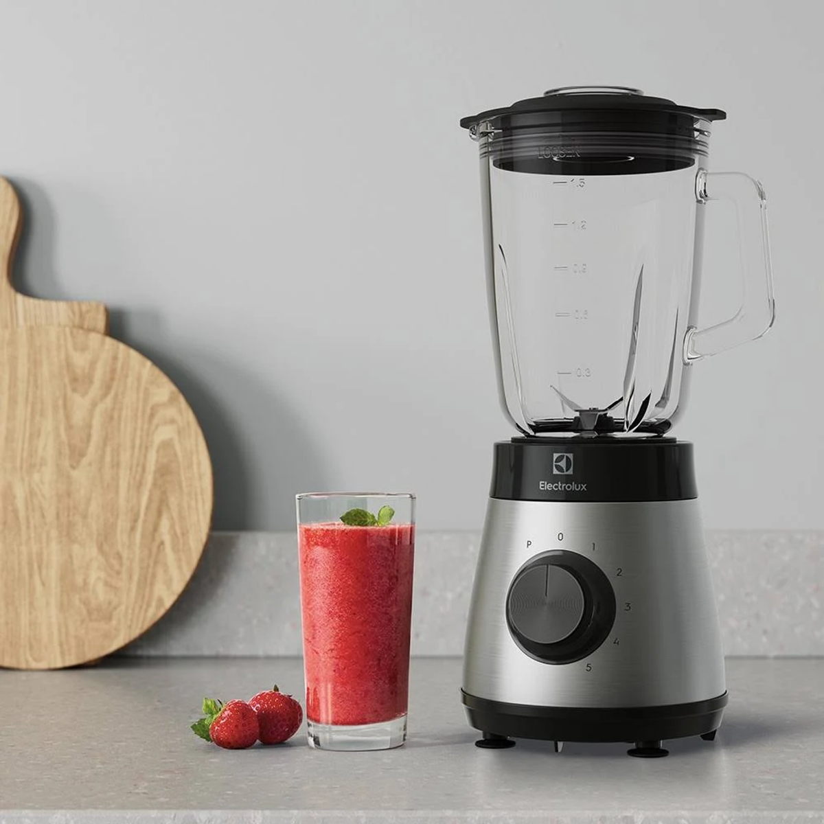 Una licuadora con un vaso de vidrio sobre una base plateada, un vaso de batido rojo y fresas.