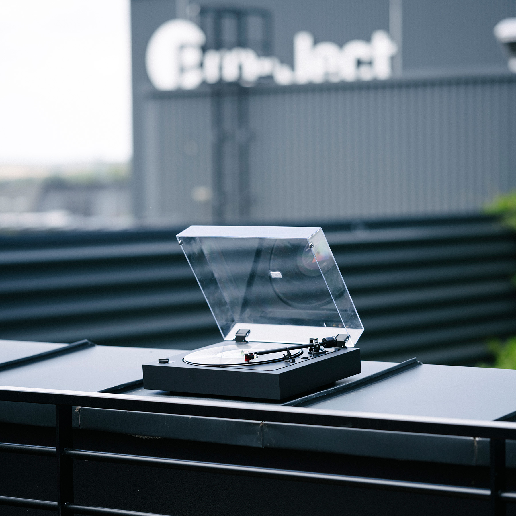 Czarny gramofon z przezroczystą pokrywą, z budynkiem w tle.