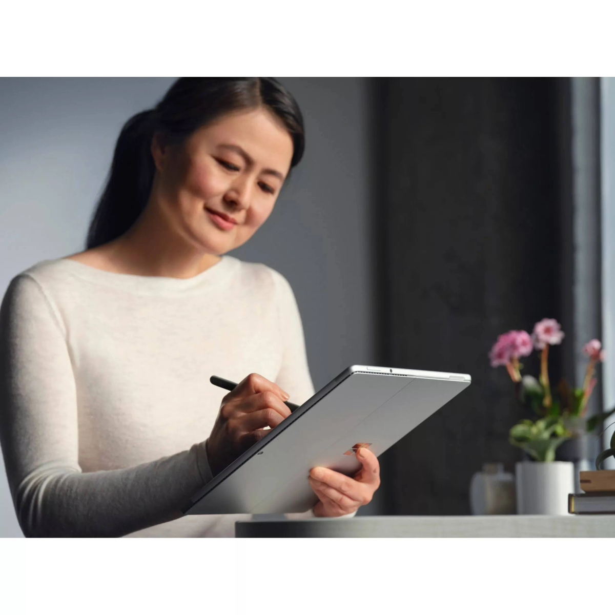 Eine lächelnde Frau benutzt einen Stift auf einem Tablet. Im Hintergrund sind Blumen und Bücher.