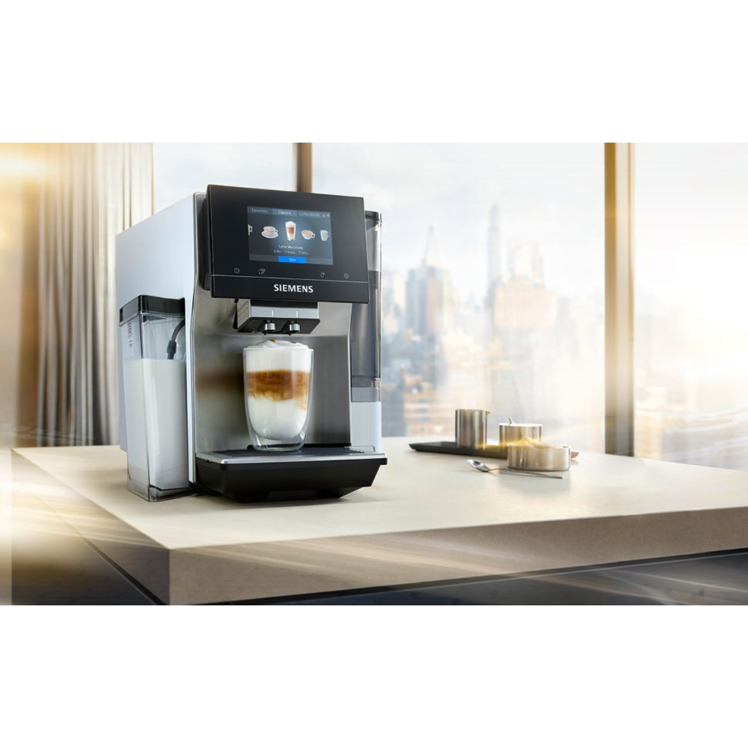 Una macchina da caffè Siemens argento e nera con un bicchiere di latte, davanti a una finestra.