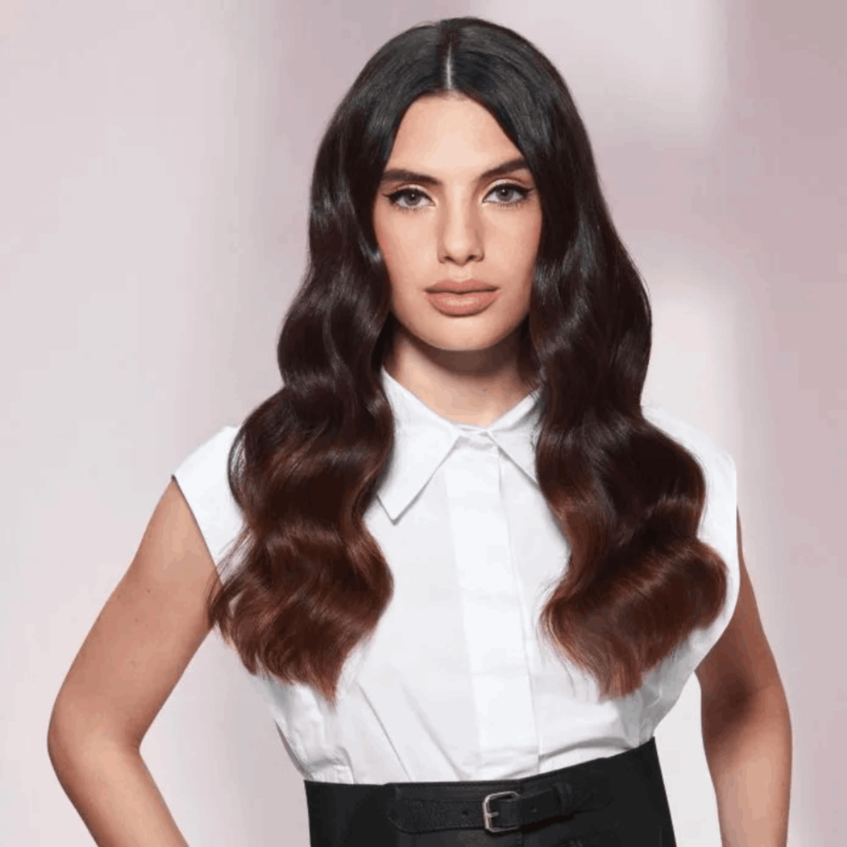 Kobieta z długimi, falowanymi brązowymi włosami, w białej koszuli i czarnej spódnicy. Patrzy w kamerę.