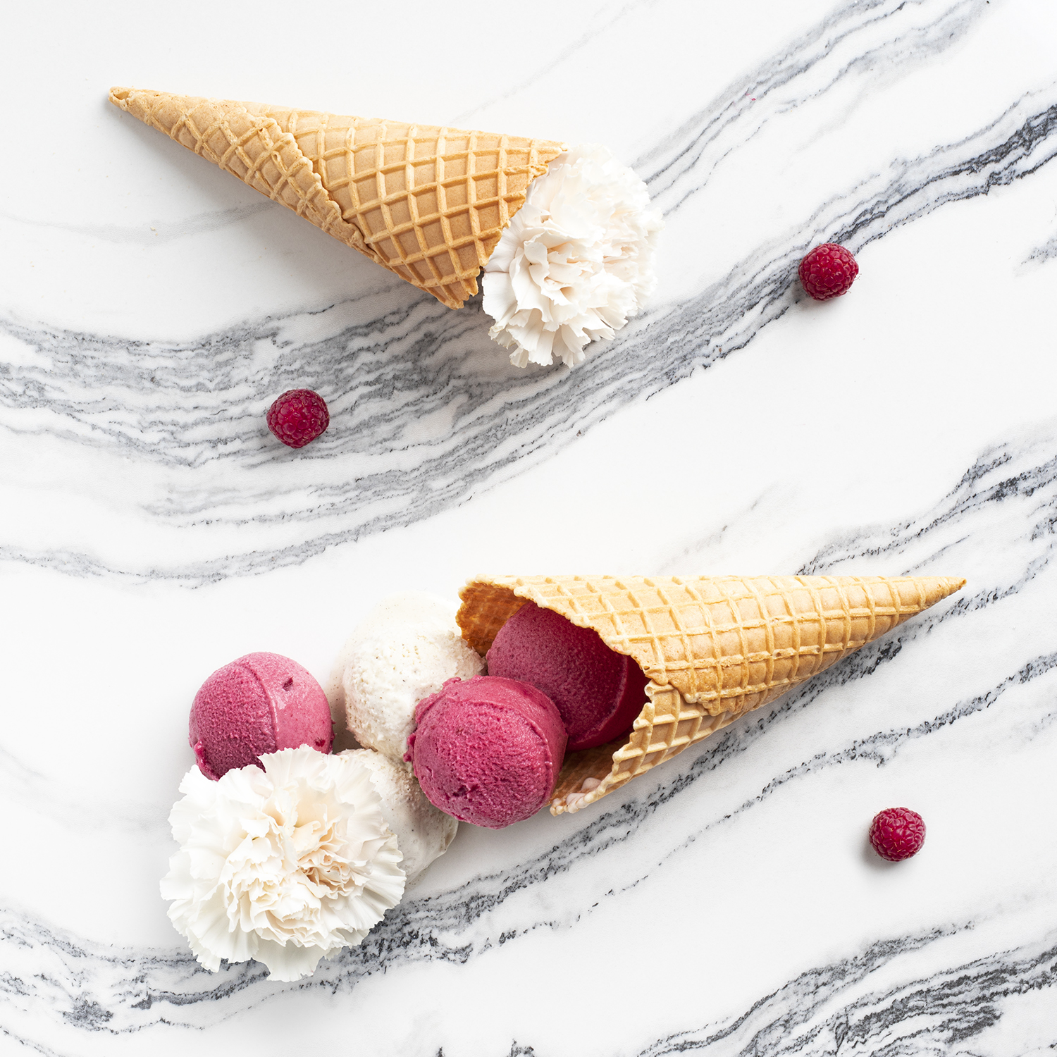 Dwa rożki waflowe wysypują gałki lodów i kwiaty na marmurową powierzchnię, z rozsianymi malinami.