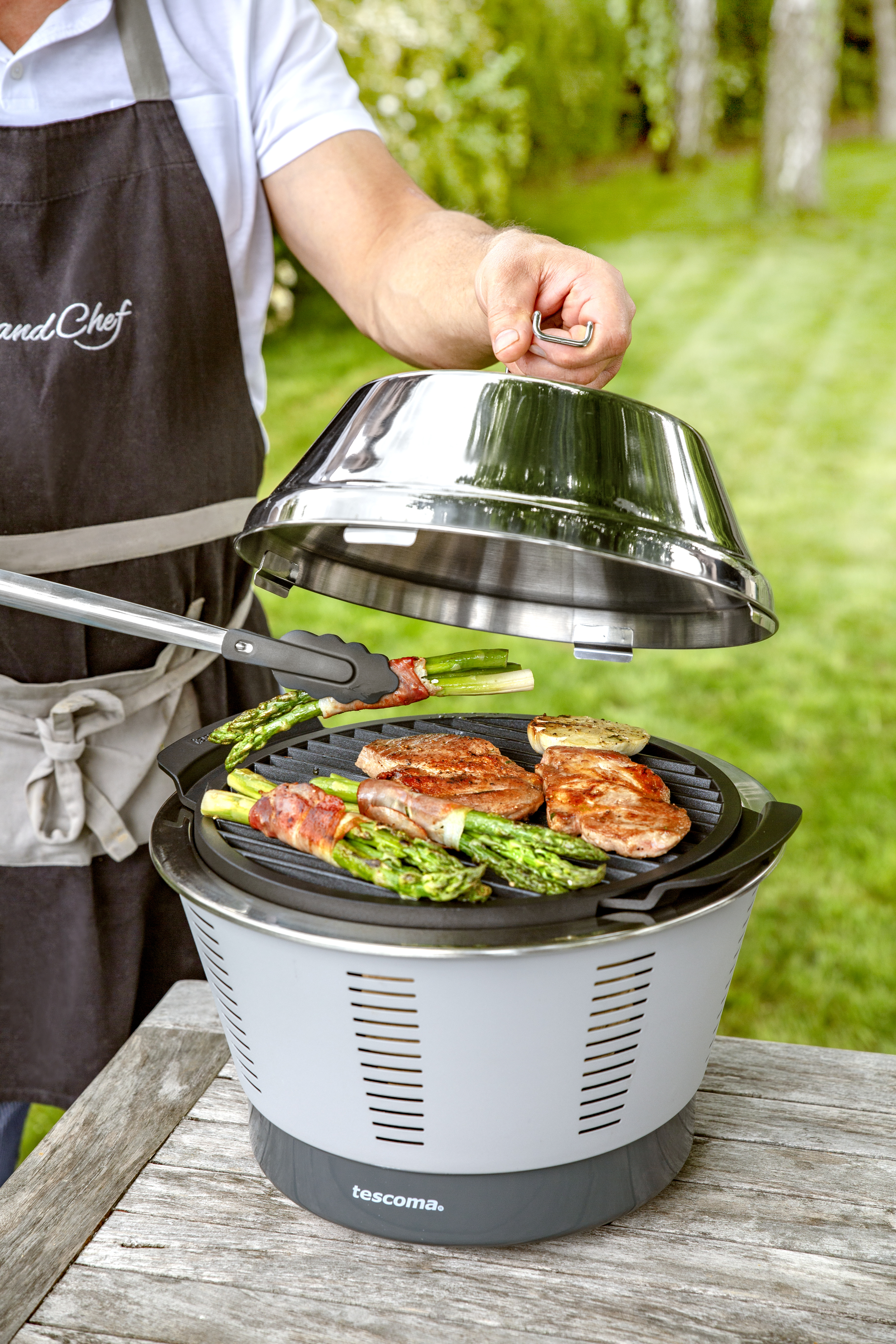 Eine Person grillt Essen auf einem Grill. Der Grill ist silber und weiß. Die Person trägt eine Schürze.