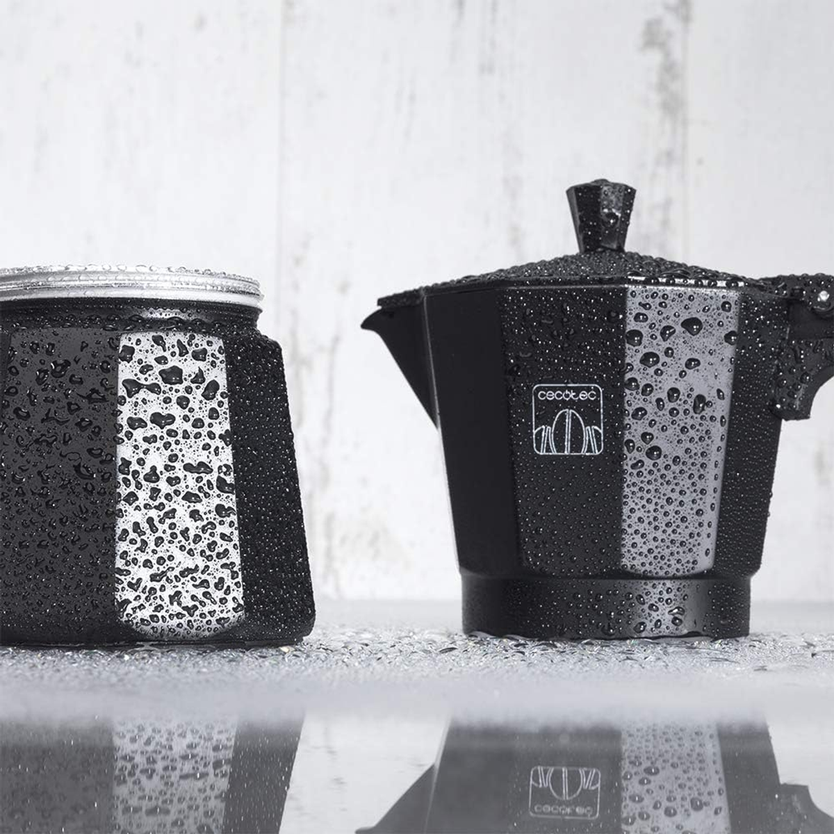 Dos cafeteras negras cubiertas de gotas de agua. Están sobre una superficie reflectante.