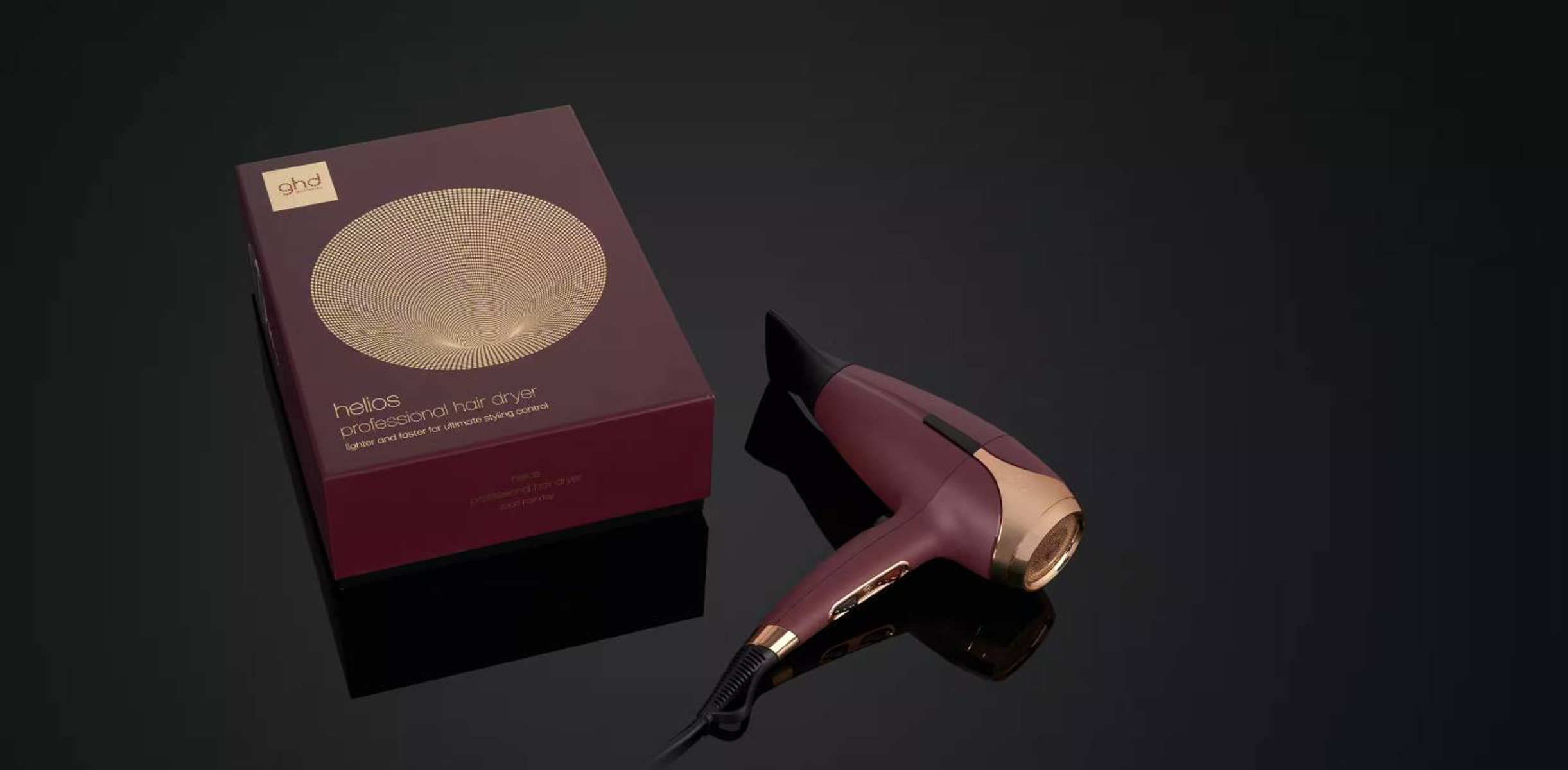 Un asciugacapelli bordeaux e una scatola con un design a cerchio dorato sono su una superficie nera.