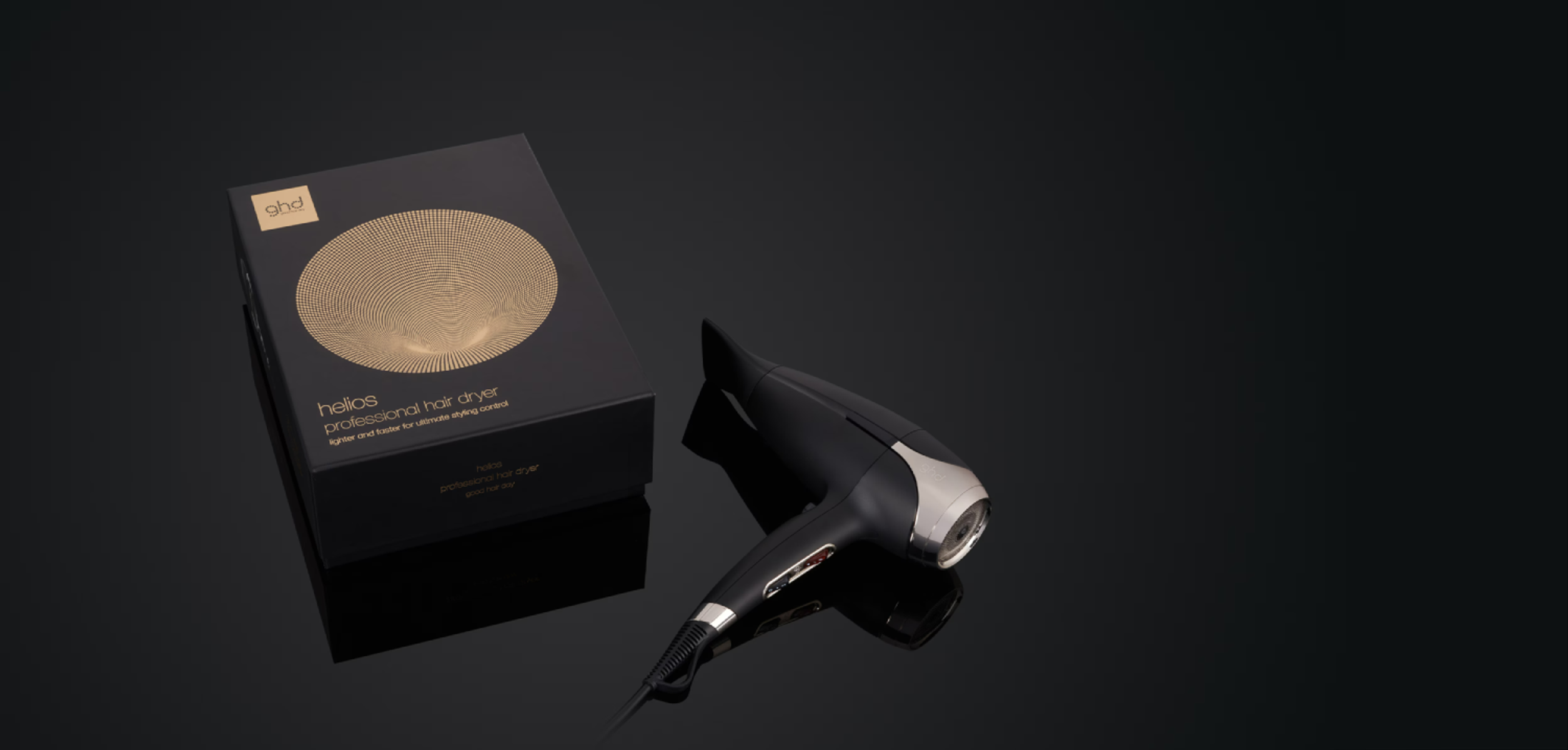 Secador de pelo y caja ghd negros sobre una superficie negra reflectante. La caja tiene un diseño dorado.