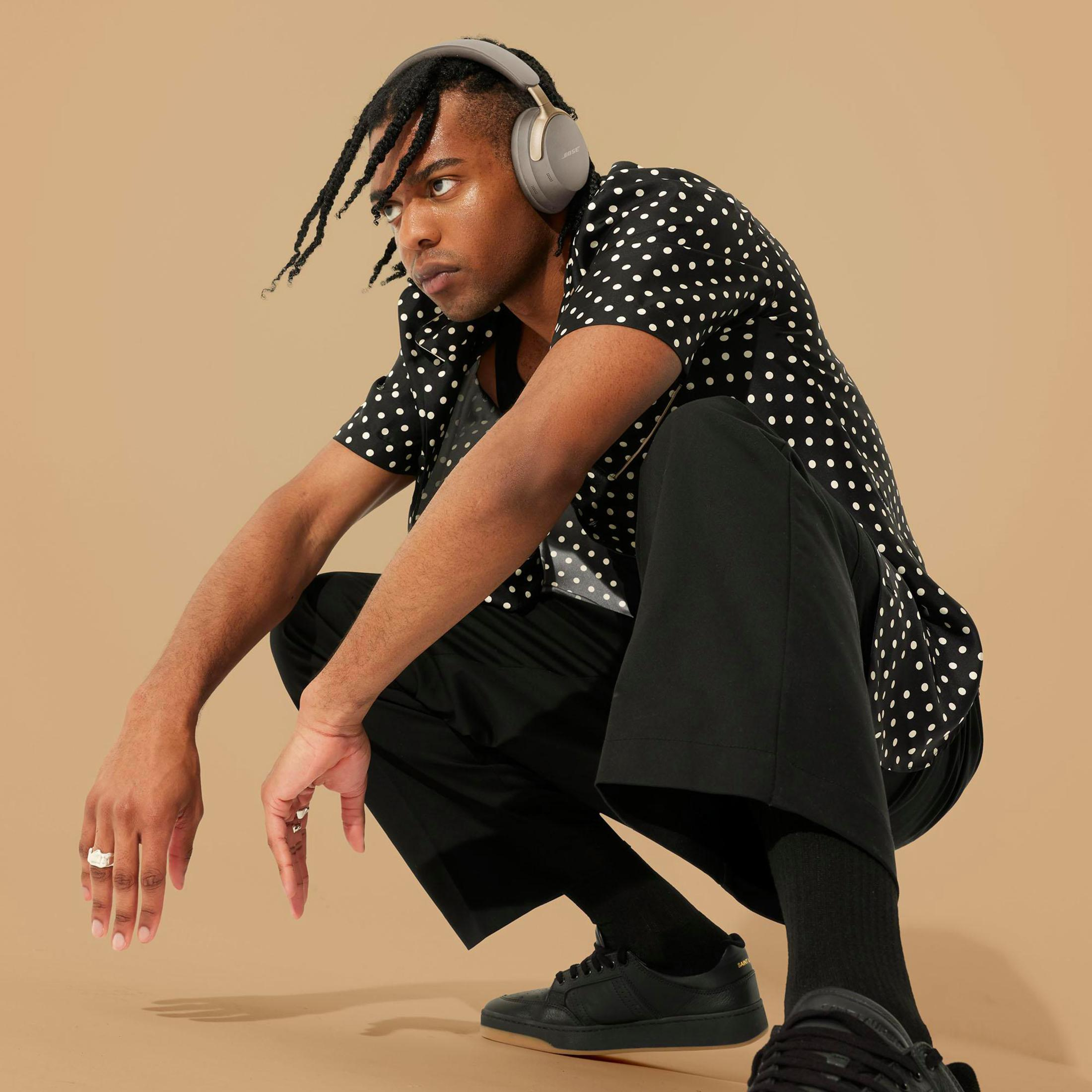 Czarny mężczyzna kuca w szarych słuchawkach, koszuli w kropki i czarnych spodniach. Beżowe tło.