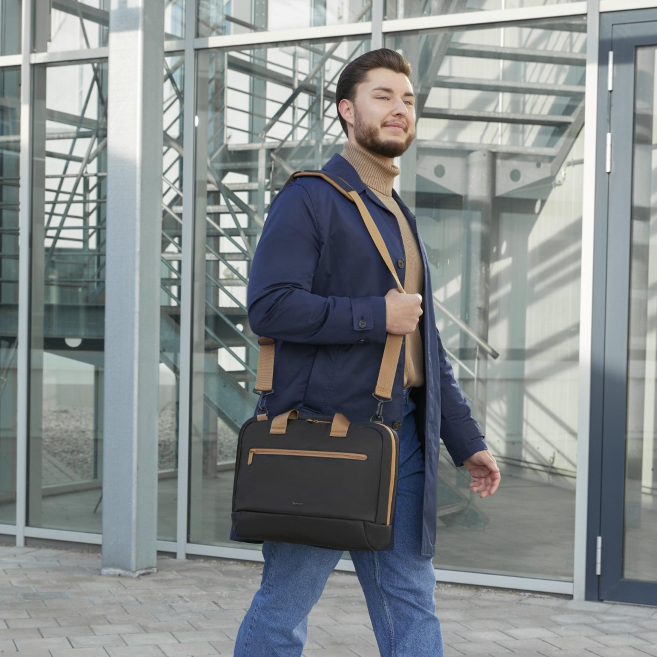 Mężczyzna w niebieskim płaszczu niosący czarną torbę z brązowym paskiem, idący na zewnątrz budynku.