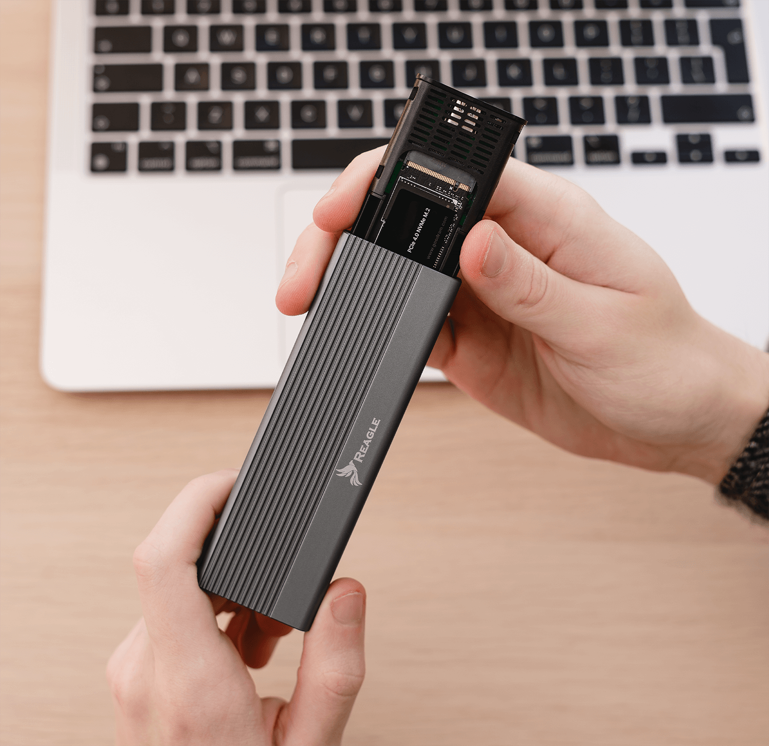 Dłonie trzymające szarą obudowę zewnętrznego dysku twardego. Laptop jest widoczny w tle.